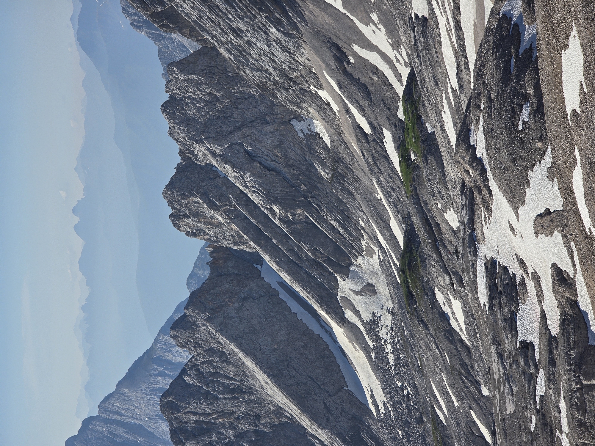 This is a high-angle, telephoto shot of the Zugspitze mountain range, near Garmisch-Partenkirchen, Germany. The image captures the rugged, grey, rocky slopes of the mountains, streaked with patches of white snow. The terrain is steep and appears to be a natural, undeveloped landscape. In the foreground, the rocky ground is interspersed with melting snow and ice. Further up the slopes, the rock faces are deeply grooved and fractured, evidence of weathering and geological activity. Patches of green vegetation are visible in a few areas on the lower slopes. In the background, a series of distant mountain ranges fade into a hazy, light blue sky, suggesting a vast and expansive mountainous region. The scene is devoid of any man-made structures or human presence, emphasizing the raw, natural beauty of the alpine environment.