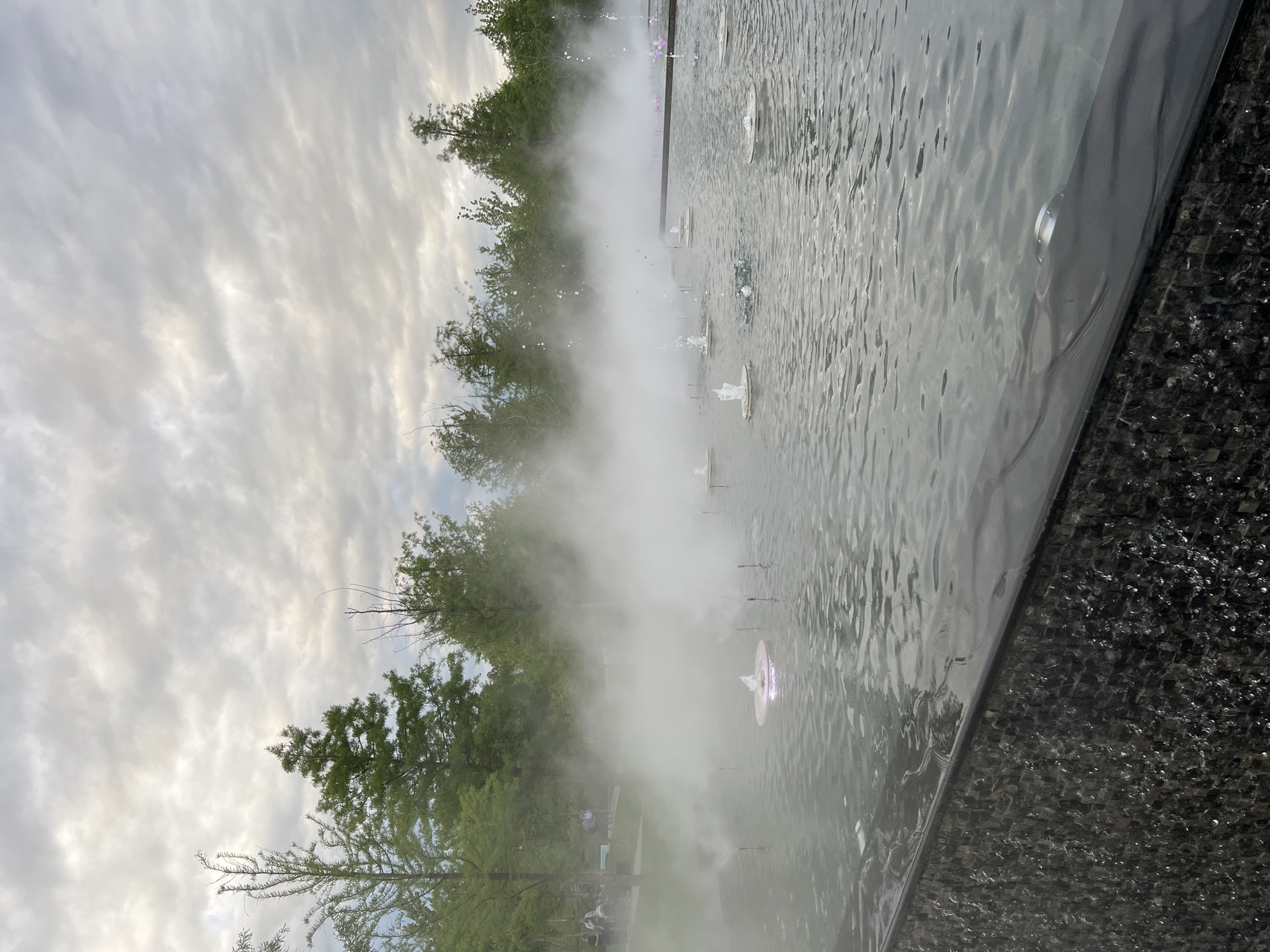 The image depicts a water feature, possibly a fountain, in a park-like setting. The presence of mist or fog partially obscures the view, revealing trees and a cloudy sky in the background. Jets of water rise from the fountain’s surface, creating small ripples on the water. A dark tiled edge is visible on the right side of the image, suggesting the boundary of the water feature. A few people can be distinguished, standing at distance in the mist. While it is possible that the location is in Krasnodar, Russia, based on the user request, it is impossible to verify it from the visual evidence of this single image.