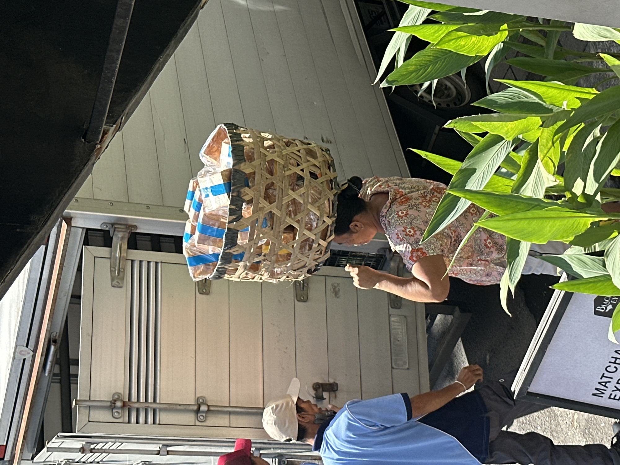 An outdoor scene in Ubud District, Indonesia, shows three people in front of a truck. In the foreground, a person wearing a light blue shirt and a beige baseball cap is facing a woman in a floral print shirt who is carrying a large woven basket filled with packaged food items on her head. The woman is looking to her left and has her left hand raised. A third person, partially visible on the left, is wearing a red cap. The truck behind them has a white wooden panelled exterior with silver accents. In the lower foreground, a sign with the words "MATCHA EXP" is partially visible. Lush green plants are in the right foreground, obscuring part of the scene. The ground appears to be paved.