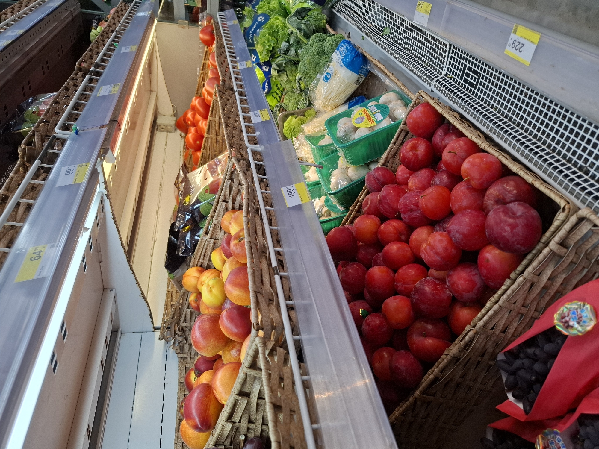 This image depicts a grocery store aisle, likely in Slyudyanka, Russia, showcasing a variety of fresh produce displayed on tiered shelving. The shelves are constructed of metal and wood, with white wire grids forming the base of each display.
In the foreground, a woven basket is filled with ripe, red plums. To the left of the plums, a red-ribboned container holds a cluster of dark purple grapes.
The middle shelf displays a variety of vegetables. Green plastic containers hold mushrooms. Behind the mushrooms are bunches of broccoli and heads of lettuce. A price tag labeled "219" is visible on the shelf edge.
The top shelf features a woven basket overflowing with peaches and nectarines, a mix of red and yellow hues. Further back, a basket is filled with bright red tomatoes, and next to them, other produce, possibly more tomatoes or other fruits, are visible. Price tags with numbers like "64" and "99" are affixed to the shelf edges.
The overall environment suggests a well-stocked grocery store with a focus on fresh produce. The lighting appears to be from overhead fluorescent fixtures, illuminating the colorful fruits and vegetables.