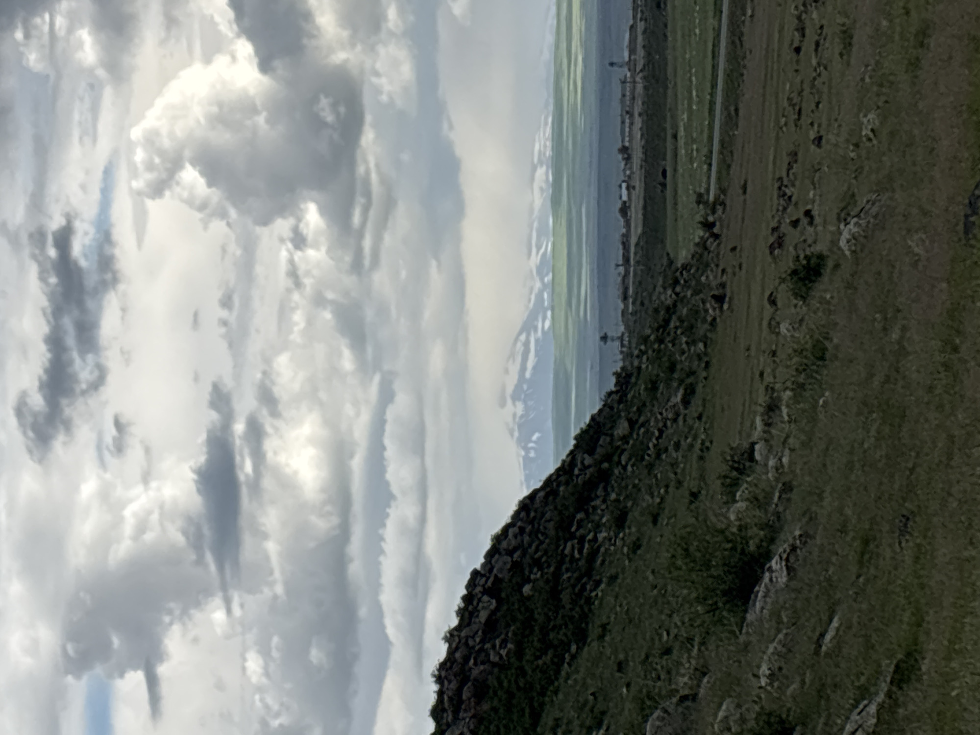 The image shows a landscape in the vicinity of Tlik, Armenia. The foreground features a rocky, grassy hillside, sloping upwards from right to left. In the midground, there is a view of cultivated fields and distant buildings or structures under a sky with prominent clouds. There is no visible human presence or interaction in the scene.