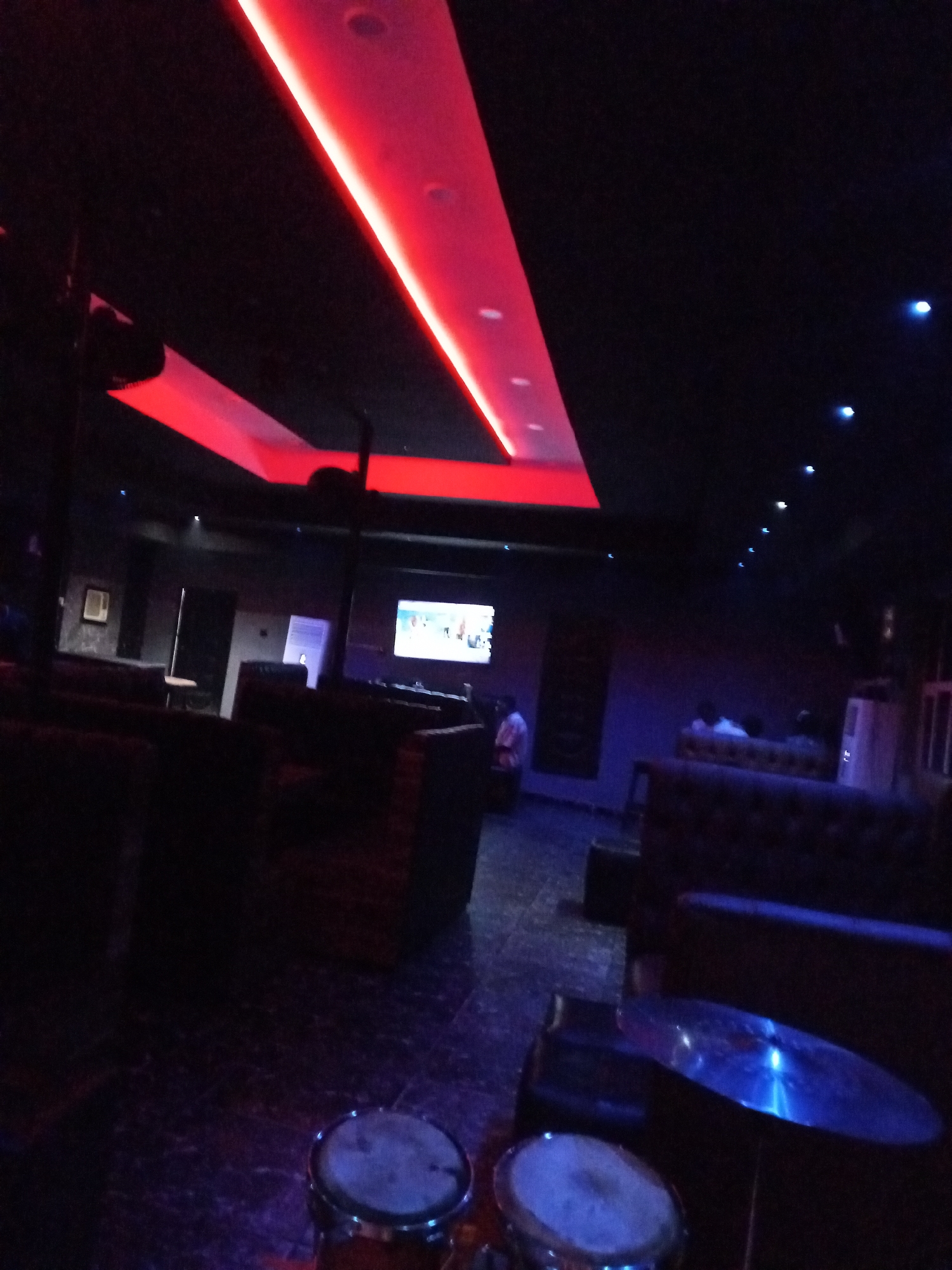The image depicts the interior of a dimly lit establishment, possibly a lounge or bar, in Lokoja, Nigeria. Red neon lights illuminate the ceiling in a dramatic fashion, contrasting with the dark ambiance. The floor appears to be tiled and reflects the ambient lighting, giving it a glossy, almost wet appearance.
In the foreground, the lower part of the image shows two drum-like objects, one with a cymbal positioned above it, suggesting a musical setup within the venue. The objects are partially obscured by the darkness, but their metallic sheen is visible.
Further into the scene, plush seating arrangements, likely couches or booths, are visible. These are upholstered in dark materials, blending into the shadows. A large screen displaying an image is mounted on the wall in the mid-ground, and a few individuals are dimly discernible in the background, seated or standing, engaged in various activities within the establishment. The overall impression is one of a contemporary entertainment space, designed for nightlife or social gatherings.