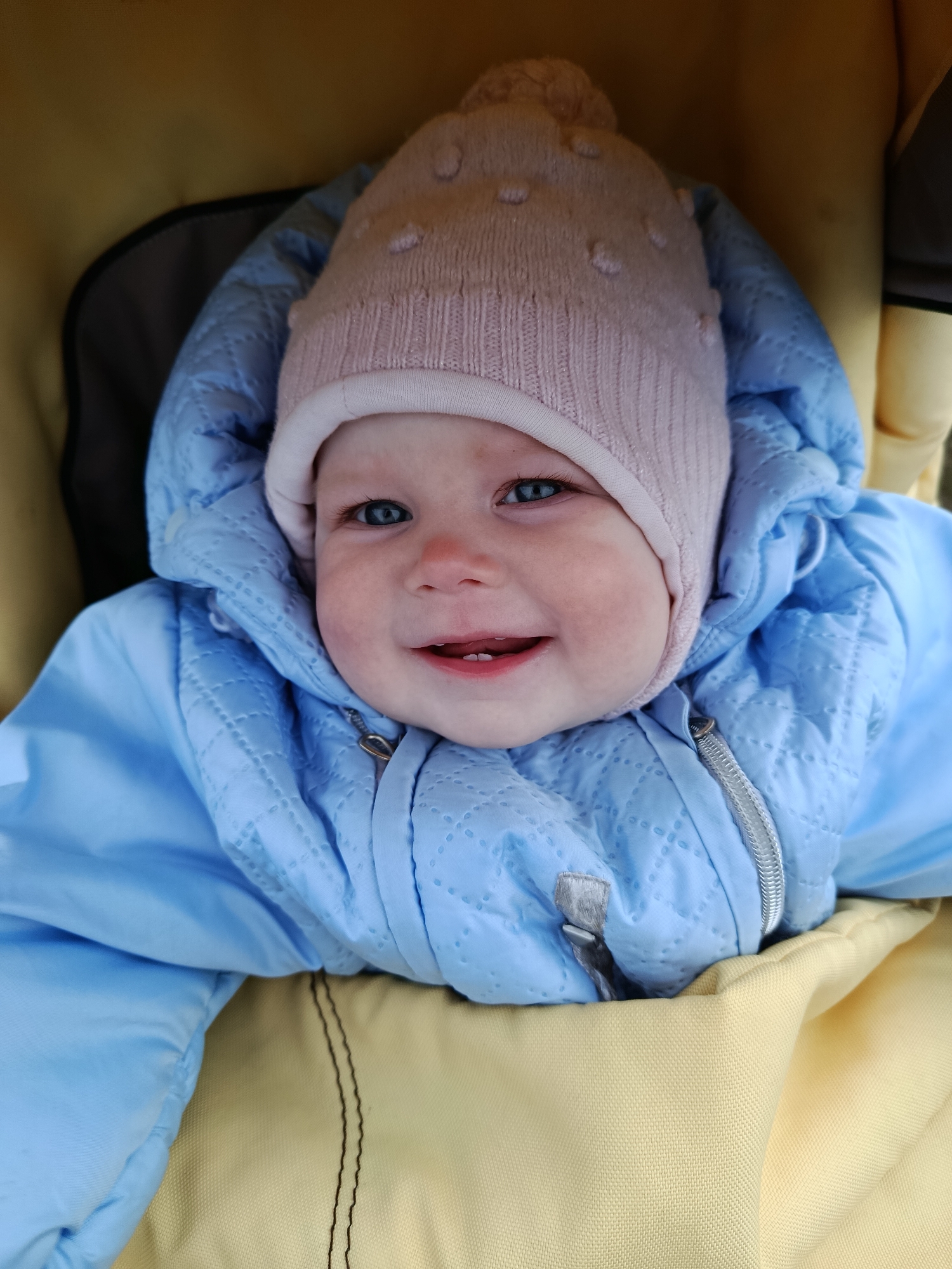 A close-up, eye-level shot shows a smiling baby bundled in warm clothing, looking directly at the camera. The baby's face is central to the frame, revealing bright blue eyes and rosy cheeks. A wide, joyful smile displays two small lower teeth.
The baby is wearing a light pink knitted hat with a textured pattern and small, evenly spaced bobbles. Below the hat, the baby is dressed in a light blue quilted jacket or snowsuit, featuring a diamond stitching pattern. A silver-colored zipper is visible down the front of the jacket. The hood of the jacket is pulled up, framing the baby's face.
The baby appears to be nestled within a stroller or car seat, which is lined with a muted yellow or tan fabric that fills most of the background. Some darker grey or black structural elements of the carrier are visible on the left side.
The warm attire suggests a cool or cold environment. The baby appears happy and content. No specific location cues, time of day, or visible text are present in the image.
