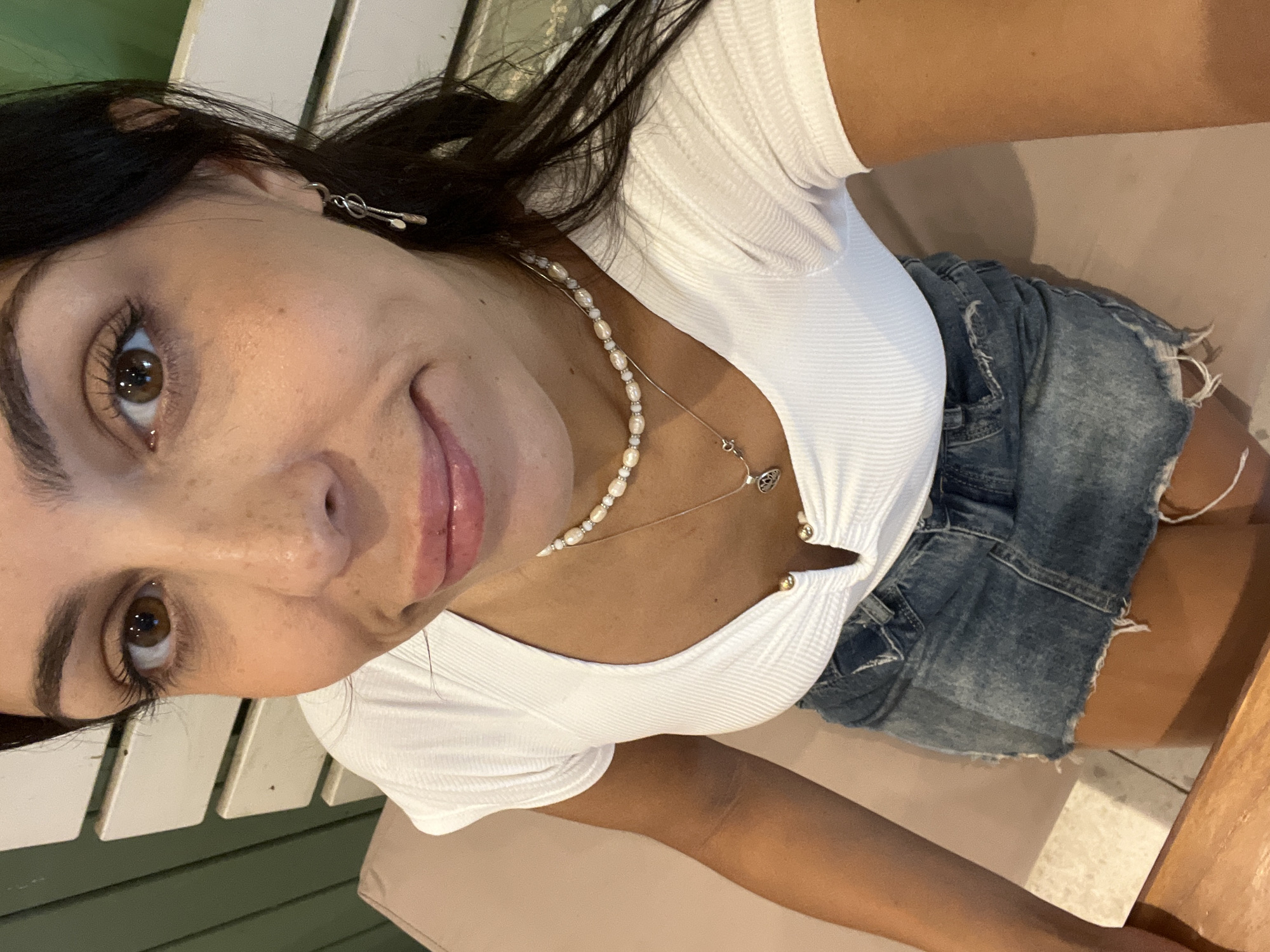 A tanned-skinned female is in the foreground, appearing to take a selfie. She has dark hair, brown eyes, and freckles. She is wearing a white, ribbed short-sleeved shirt with buttons at the neckline, a pearl necklace, and a silver necklace with a pendant. She is also wearing a denim mini skirt. She is seated on what appears to be a white slat bench. The background includes what looks like a green slat surface and a tan-colored chair or bench. The location is Kuta Selatan, Indonesia.