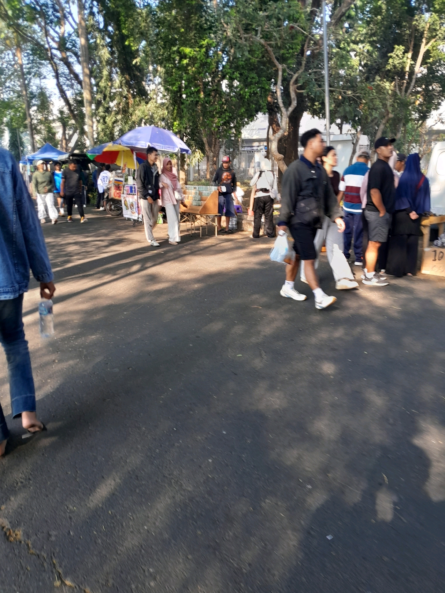 The image depicts a street scene in Kedungpedaringan, Indonesia, characterized by a gathering of people and various stalls under trees. The foreground shows a paved road with shadows cast by overhead foliage. A person wearing a denim jacket and blue jeans is walking towards the left, holding a water bottle. In the mid-ground, several individuals are seen walking and standing around market stalls. One stall has colorful umbrellas and appears to be selling food. Another stall on the right has a wooden structure with a product labeled "10". People are dressed in casual attire, with some wearing headscarves. The overall environment suggests a public space, possibly a local market or a pedestrian area.