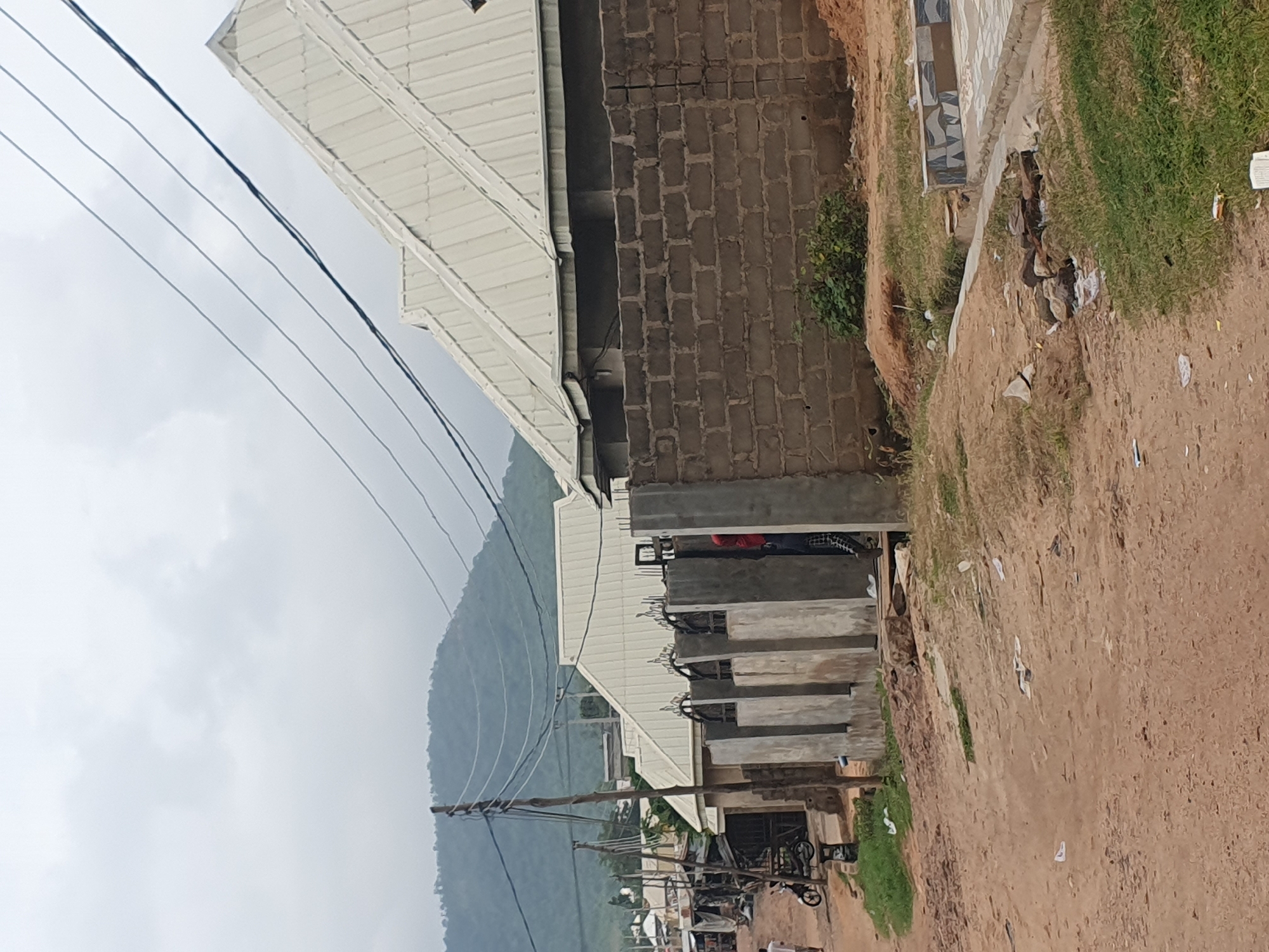 The image captures a street scene in Lokoja, Nigeria, under an overcast sky. In the foreground, a dusty, unpaved road stretches into the distance, with scattered pieces of white paper and some rocks. To the right, a newly constructed pathway or patio area with a decorative tiled border is visible, bordered by green grass and more dirt.
Dominating the midground are several buildings under construction, characterized by their light-colored, corrugated metal roofs. One building features a wall constructed from grey cinder blocks. Adjacent to this, a fence made of concrete pillars and ornate metalwork separates properties. A person in a red shirt and dark pants is partially visible behind this fence, appearing to be interacting with the construction.
Further down the street, a row of market stalls or shops with tarpaulin roofs lines the left side, some appearing to be open for business. A parked motorcycle is visible near one of these stalls. A person in a white shirt is walking away from the camera on the left side of the road.
In the background, a lush, green mountain range rises against the hazy sky, suggesting a natural landscape bordering the urban or semi-urban development. Utility poles and a network of electrical wires crisscross the upper portion of the image, indicating the presence of electricity infrastructure. The overall impression is one of ongoing development and daily life in a Nigerian city.