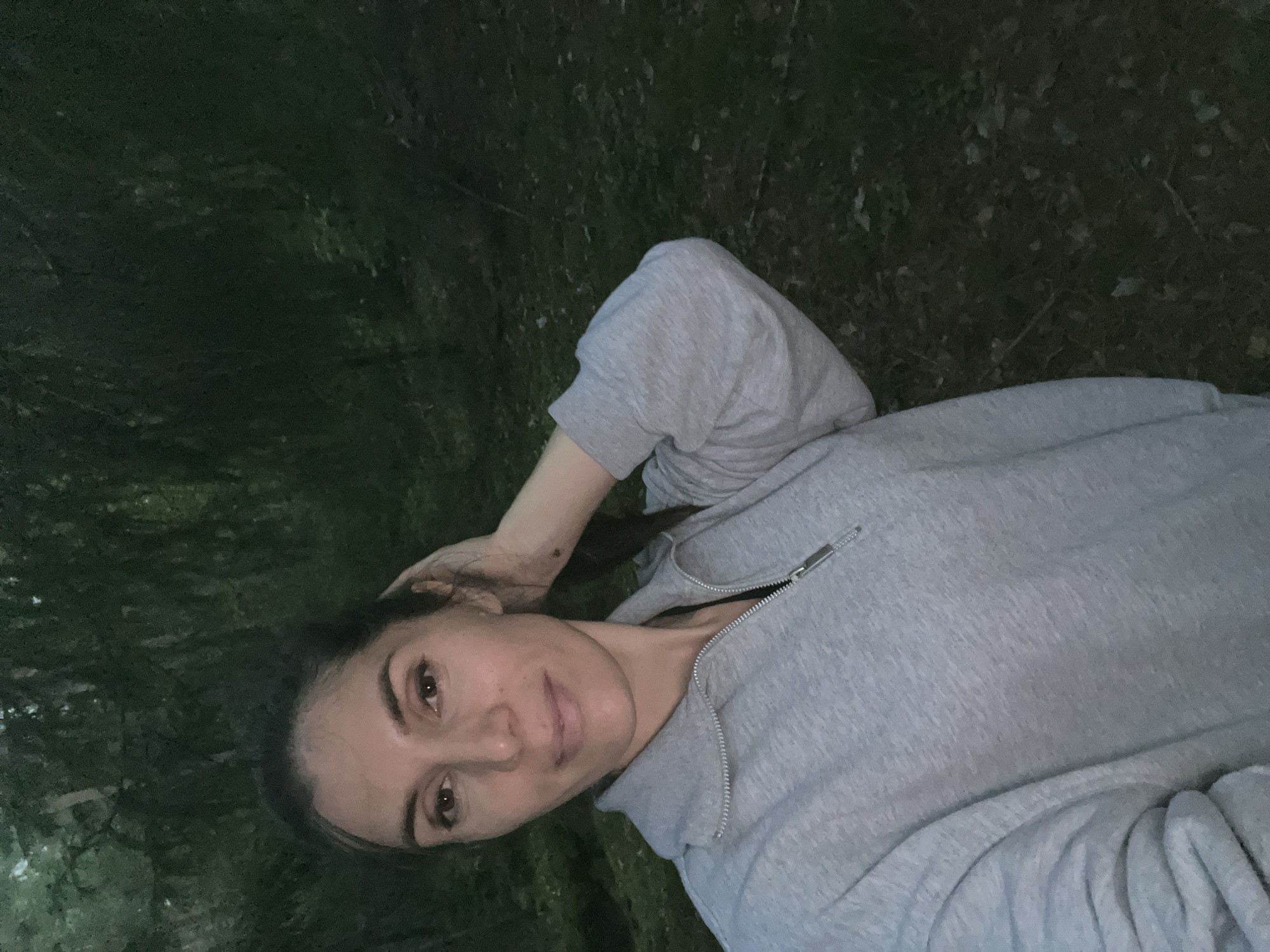 This is a low-light, self-taken photograph of a woman lying down outdoors. She is wearing a grey zip-up hoodie. Her head is propped on her arm, and her hair is pulled back. She is looking towards the camera with a relaxed expression. The background appears to be grass and foliage, suggesting a natural, outdoor setting. The time of day seems to be evening or night due to the low light conditions. The woman's face is illuminated, possibly by the camera's flash or another light source. There is no visible text in the image.