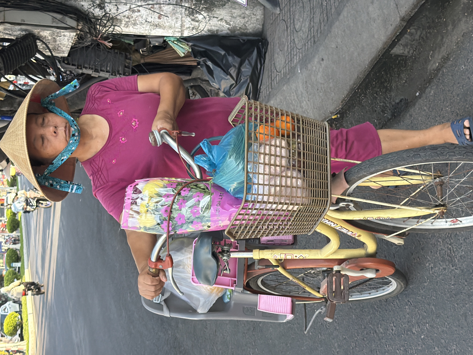 A person wearing a conical hat and a pink shirt stands next to a bicycle on a street in Bac Nha Trang Ward, Vietnam. The person's face is partially obscured by a blue cloth tied under their chin. The bicycle has a pink child seat and a basket filled with items, including what appears to be flowers in a pink bag and blue plastic bags. The street is paved with asphalt, and a concrete sidewalk is visible to the right, lined with electrical wires and conduits. In the background, there are other people on motorbikes and some greenery. The overall scene suggests a typical street view in a Vietnamese city.