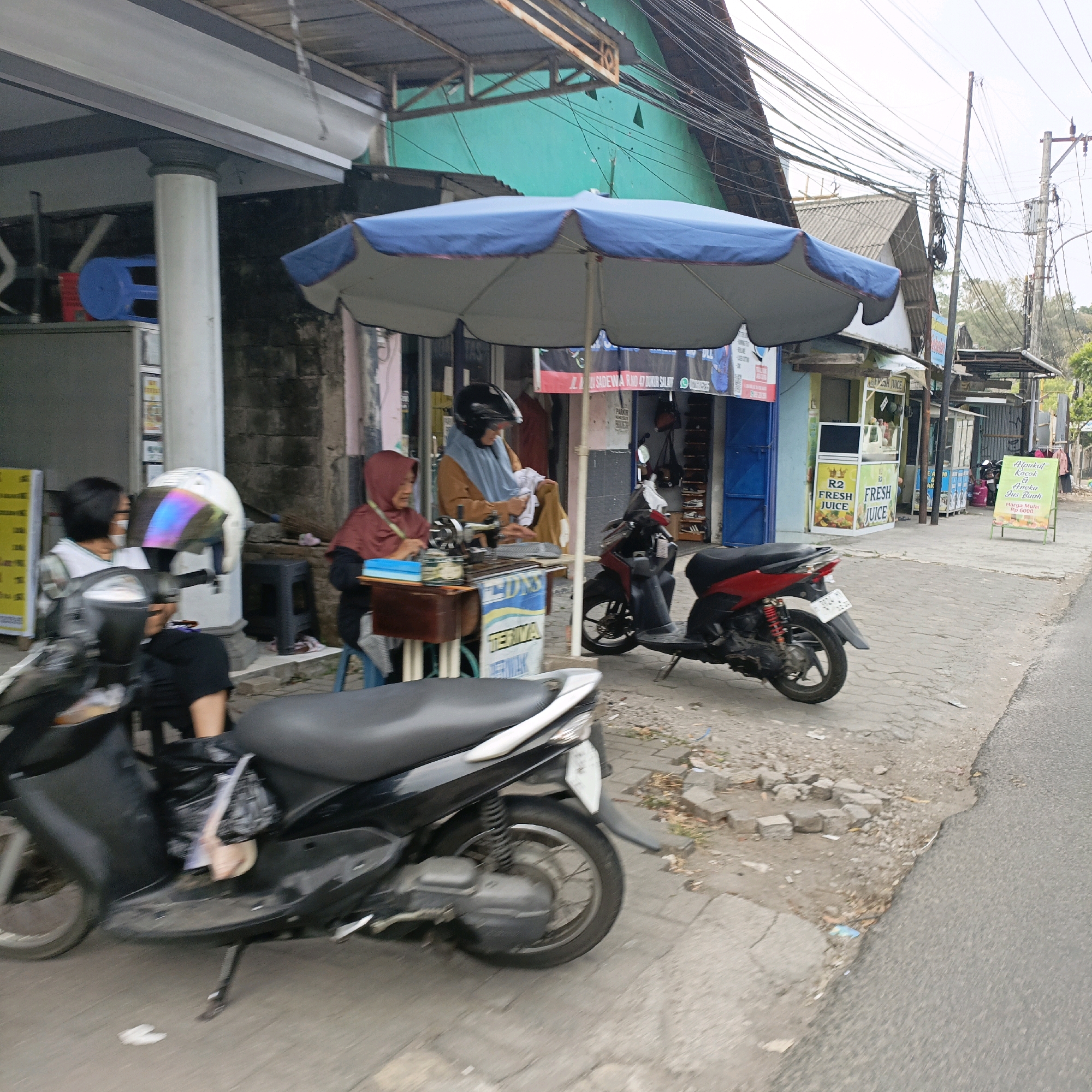 The image captures a street scene in Salatiga, Indonesia, likely a commercial area with small shops and food stalls. In the foreground, a black scooter is parked on a sidewalk, its front wheel turned slightly. To its left, a woman wearing a white face mask and a patterned shirt is seated on another scooter, wearing a white helmet with iridescent visor. She is looking towards the left. Behind her, a yellow sign is visible.
Further back, under a blue and white umbrella, a sewing station is set up. A woman wearing a reddish-brown hijab is seated on a stool, her hands resting on a sewing machine. Another woman, also wearing a reddish-brown hijab and an orange top, is seated beside her, appearing to be sewing or working with fabric. A sewing machine and various items are on a table.
To the right of the sewing station, a red and black scooter is parked. Beyond that, a row of small shops or stalls line the street. One stall has a sign with "R2 FRESH JUICE" and images of fruit. Another stall has a green sign board with text in Indonesian, advertising "Alpukat Kocok & Aneka Jus Buah" (Whipped Avocado & Various Fruit Juices), with "Harga Mulai Rp 6000" (Price Starts Rp 6000). The street itself is paved with asphalt and the sidewalk is made of concrete blocks. Overhead, a dense network of electrical wires crisscrosses the sky. The buildings in the background have various architectural styles and colors, with some showing signs of wear. The overall atmosphere suggests a bustling, everyday scene in a tropical urban environment.