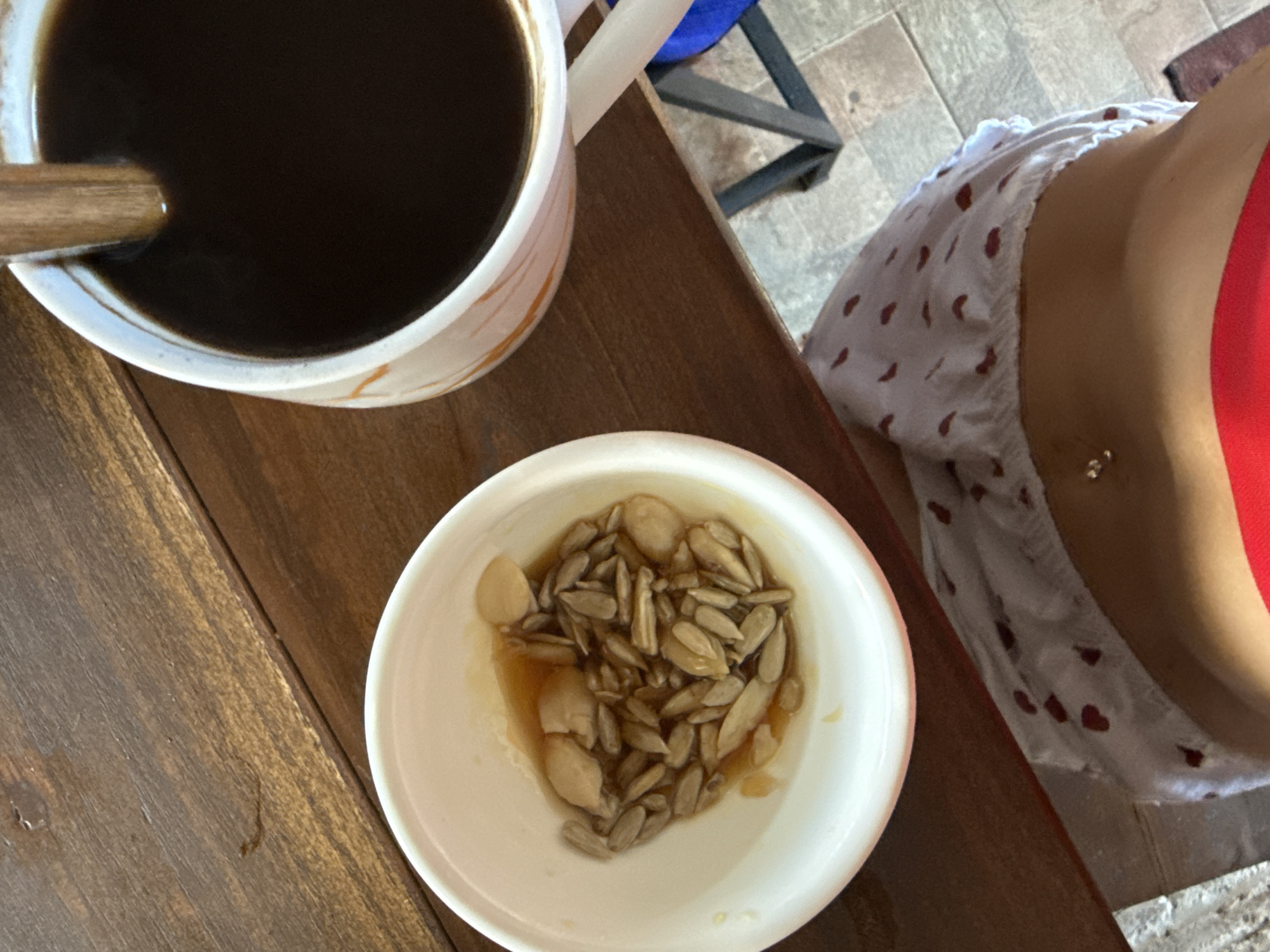 A close-up, overhead shot captures a rustic wooden table with two items: a white ceramic bowl filled with sunflower seeds and a light brown liquid, and a mug of dark coffee with a wooden stirrer. In the foreground, a person's midriff is visible, adorned with a silver belly button piercing and wearing red swimwear. The overall scene suggests a casual breakfast or snack setting in Kuta Selatan, Indonesia.