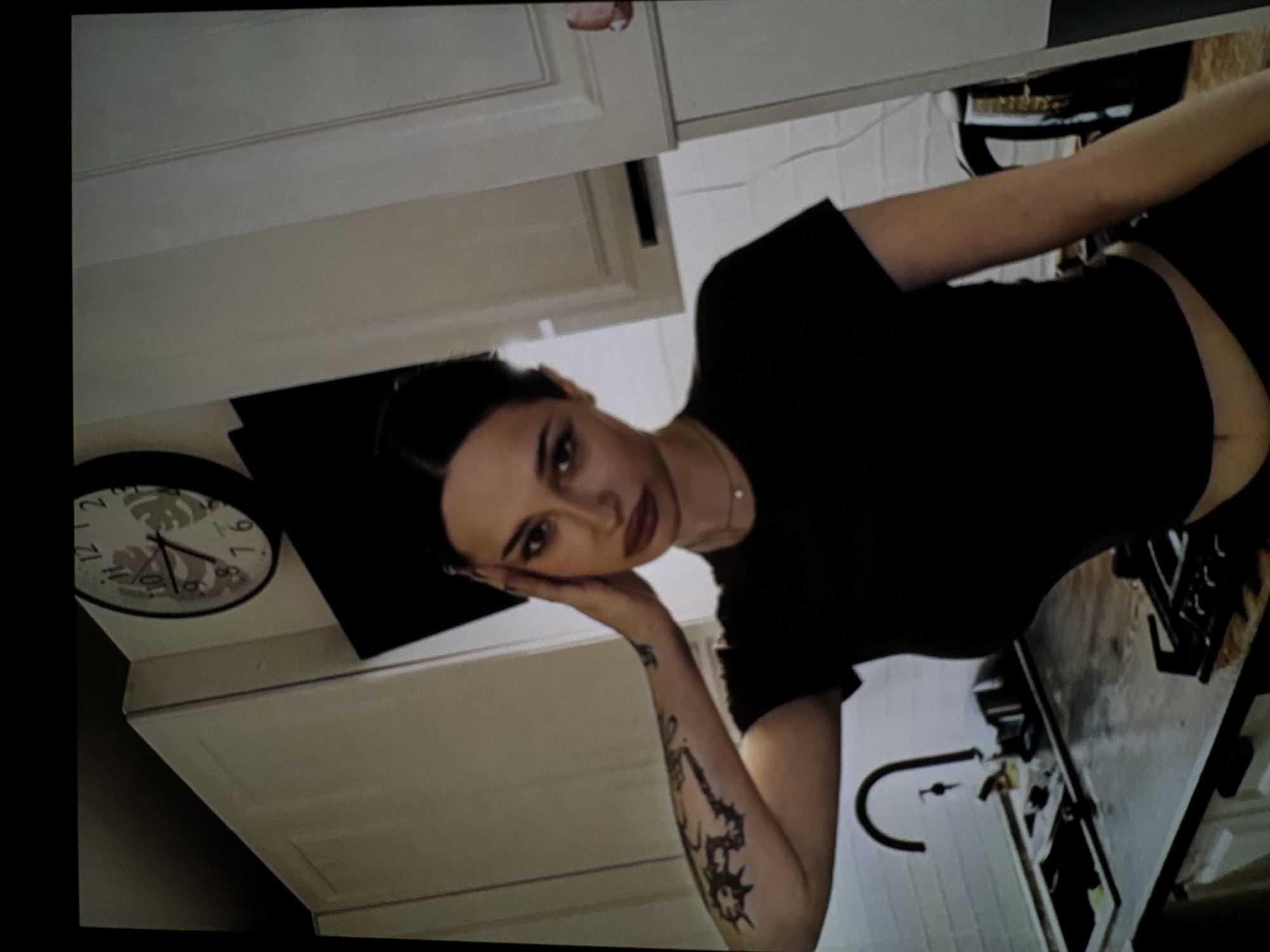 In a dimly lit kitchen, a woman with dark hair pulled back stands with her right hand on her head, her eyes looking forward. She is wearing a black t-shirt and black pants. A tattoo of what appears to be a stylized dragon is visible on her left arm. The kitchen features white cabinets, a stainless steel sink with a faucet, and a stovetop. A circular clock with Roman numerals is mounted on the wall above the cabinets. The overall impression is of a candid, indoor portrait.