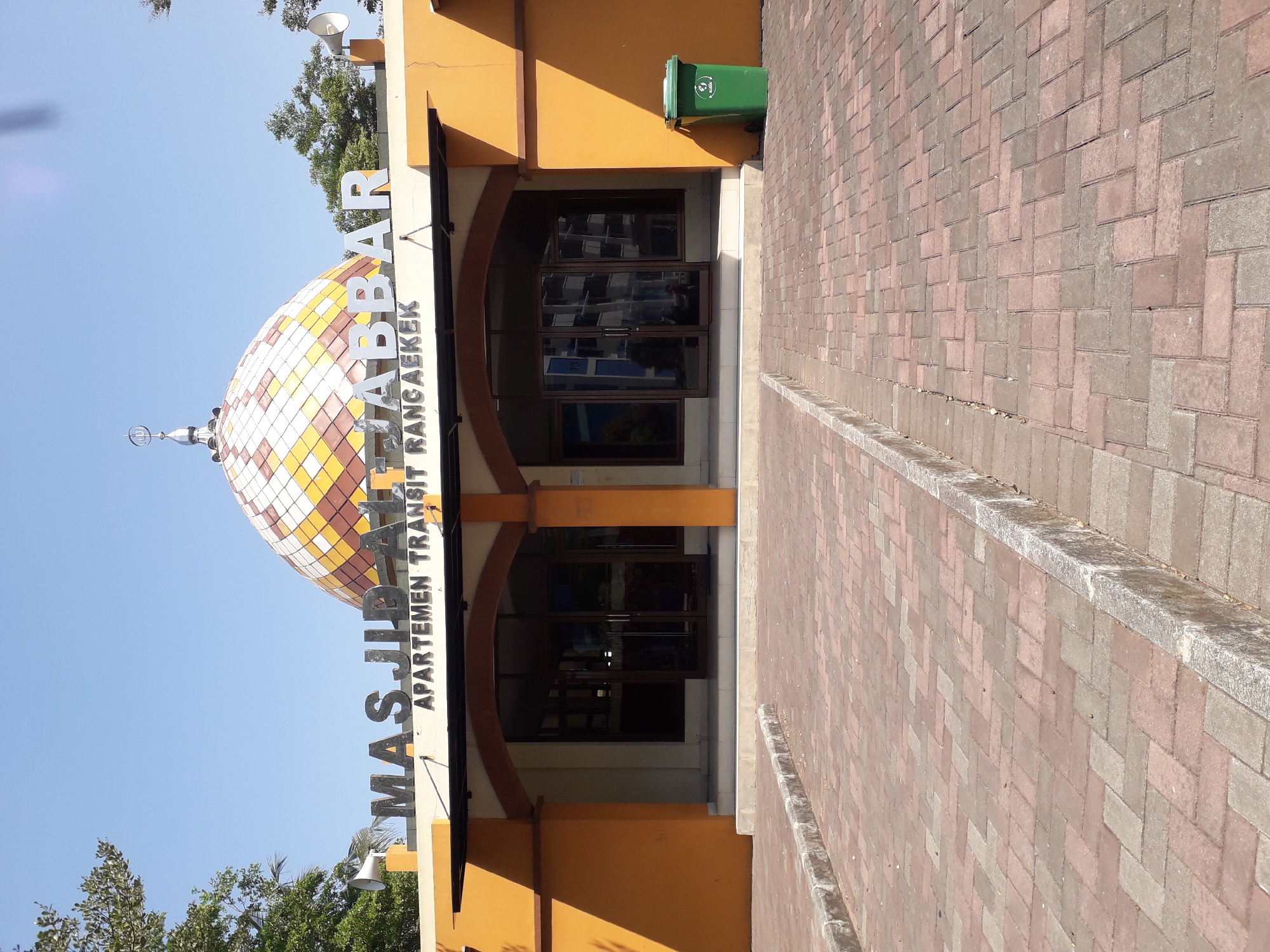 The image shows the facade of the Masjid Al-Jabbar, an apartment complex transit facility in Cankuang, Indonesia. The building has an orange exterior with a large, multi-colored tiled dome. Above the entrance, large silver letters spell out "MASJID AL-JABBAR" and "APARTEMEN TRANSIT RANGAEKKEK". The entrance features double glass doors and windows. A green trash can is visible to the right of the entrance. The foreground is a paved walkway made of red and gray bricks, with a concrete curb bordering the left side of the path. The sky is clear and blue, and trees are visible on either side of the building.