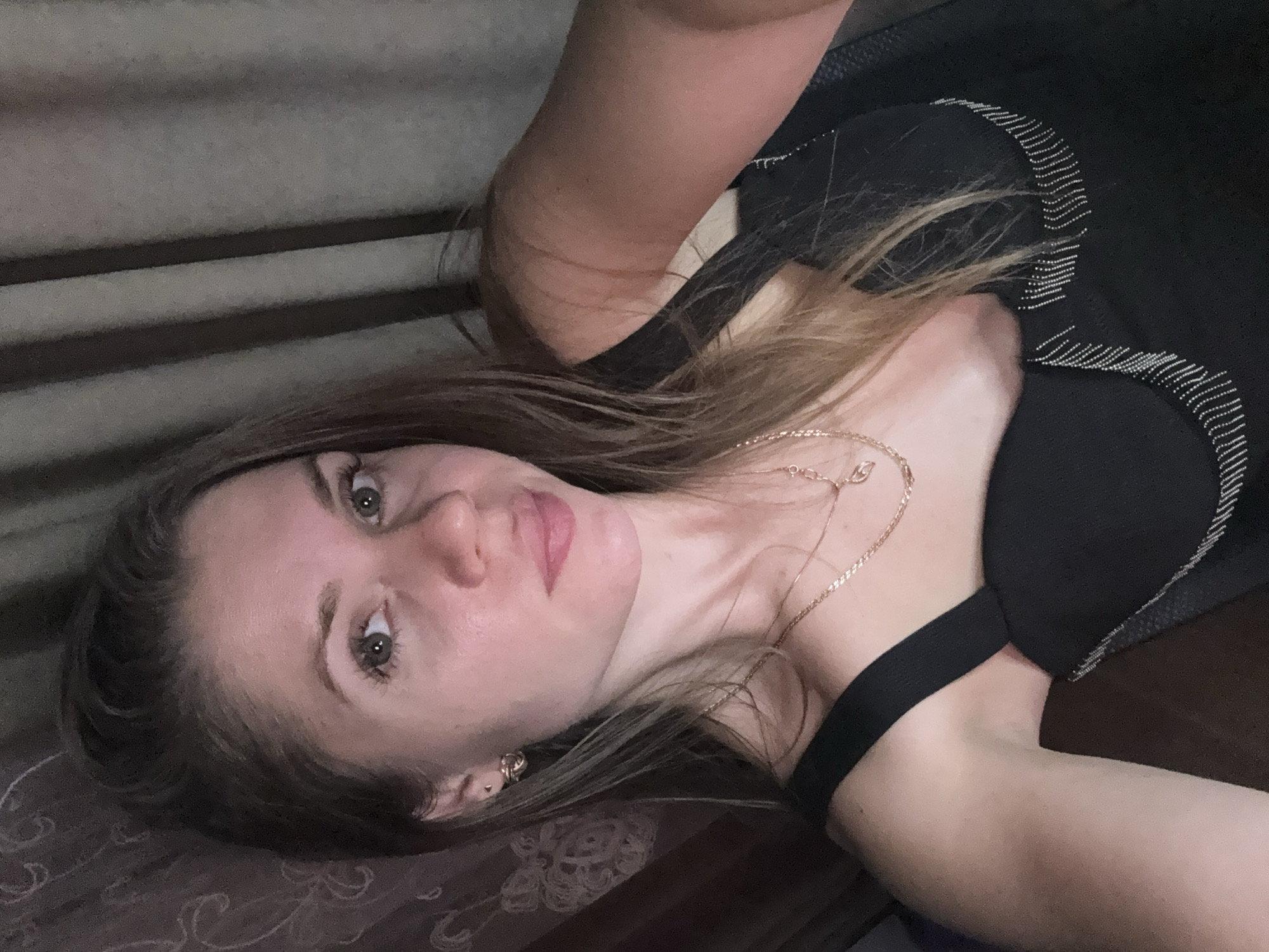 A woman is lying down, gazing at the camera. She has fair skin, light brown hair, and a gold necklace. She's wearing a black top with silver embellishments. The background shows striped wall paneling and a dark patterned surface. The location is undetermined.