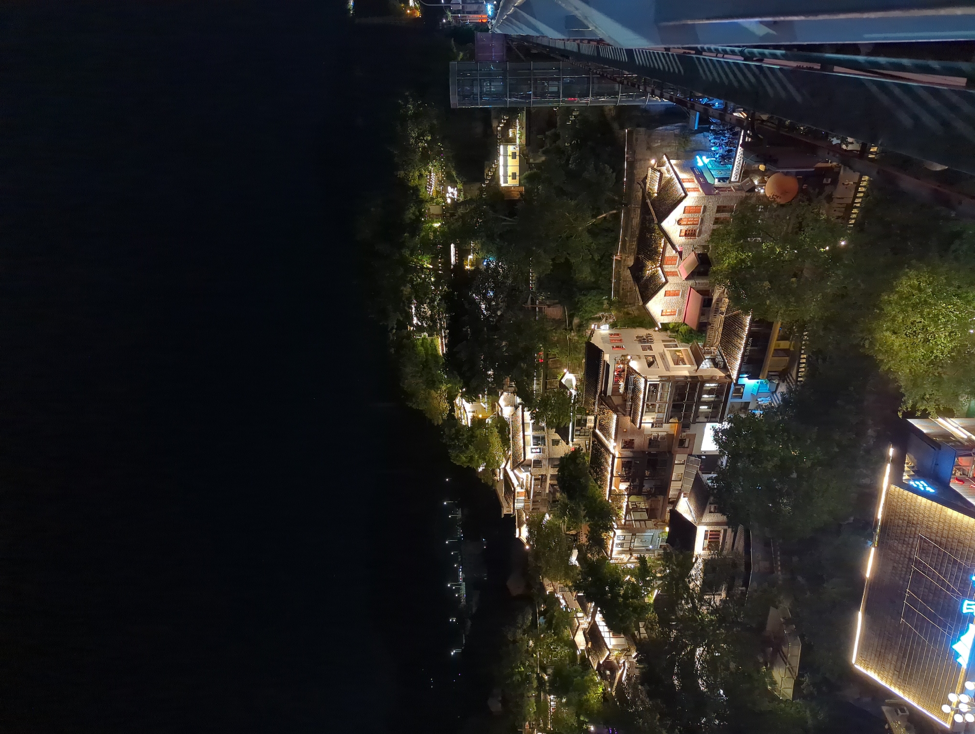 This is a nighttime aerial view of a hillside illuminated by lights, showing traditional Chinese-style buildings with tiled roofs, nestled among trees. The architecture suggests a historic or resort area. In the foreground, a modern structure with a brightly lit, multi-faceted roof and neon signage is visible. To the right, a contemporary glass elevator tower stands adjacent to a railing that extends out of the frame. In the midground and background, numerous buildings are lit, some with glowing windows and balconies, creating a warm and inviting ambiance. Trees are scattered throughout the scene, with some illuminated by spotlights. In the distance, streetlights suggest a more urban environment beyond the hillside development. A group of people are visible gathered outside a building with a blue awning.