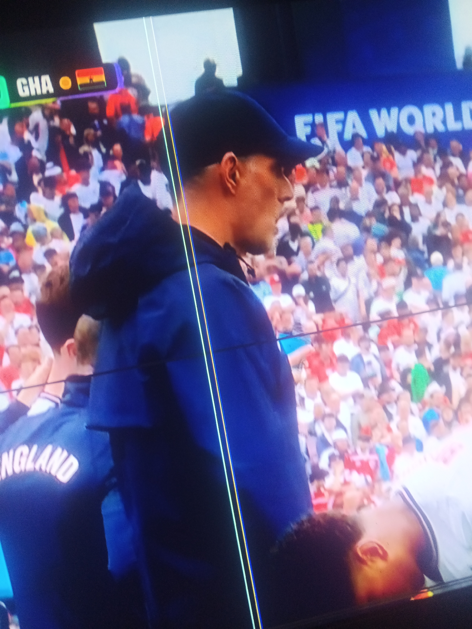 The media file captures a scene from a football match, likely during a televised broadcast. The foreground is dominated by the side profile of a man wearing a dark blue jacket and a baseball cap. His facial features are partially obscured. Behind him, a crowd of spectators is visible, suggesting a stadium setting. The crowd is a blur of colors, with many people wearing white and red attire.
To the left of the frame, a scoreboard displays "GHA" with a small flag next to it, which appears to be the flag of Ghana. Further to the right, "FIFA WORLD" is visible on a blue banner, indicating this is a FIFA World Cup event. The overall lighting suggests it is daytime. There are vertical light streaks across the image, possibly due to the capture method or screen artifacts. The presence of the "ENGLAND" text on the back of a jacket in the lower left corner implies that England is one of the teams involved in the match. The expressions of the spectators are not discernible due to the distance and blurriness. The context of Jalingo, Nigeria, places the viewing of this football match within a specific geographical location.