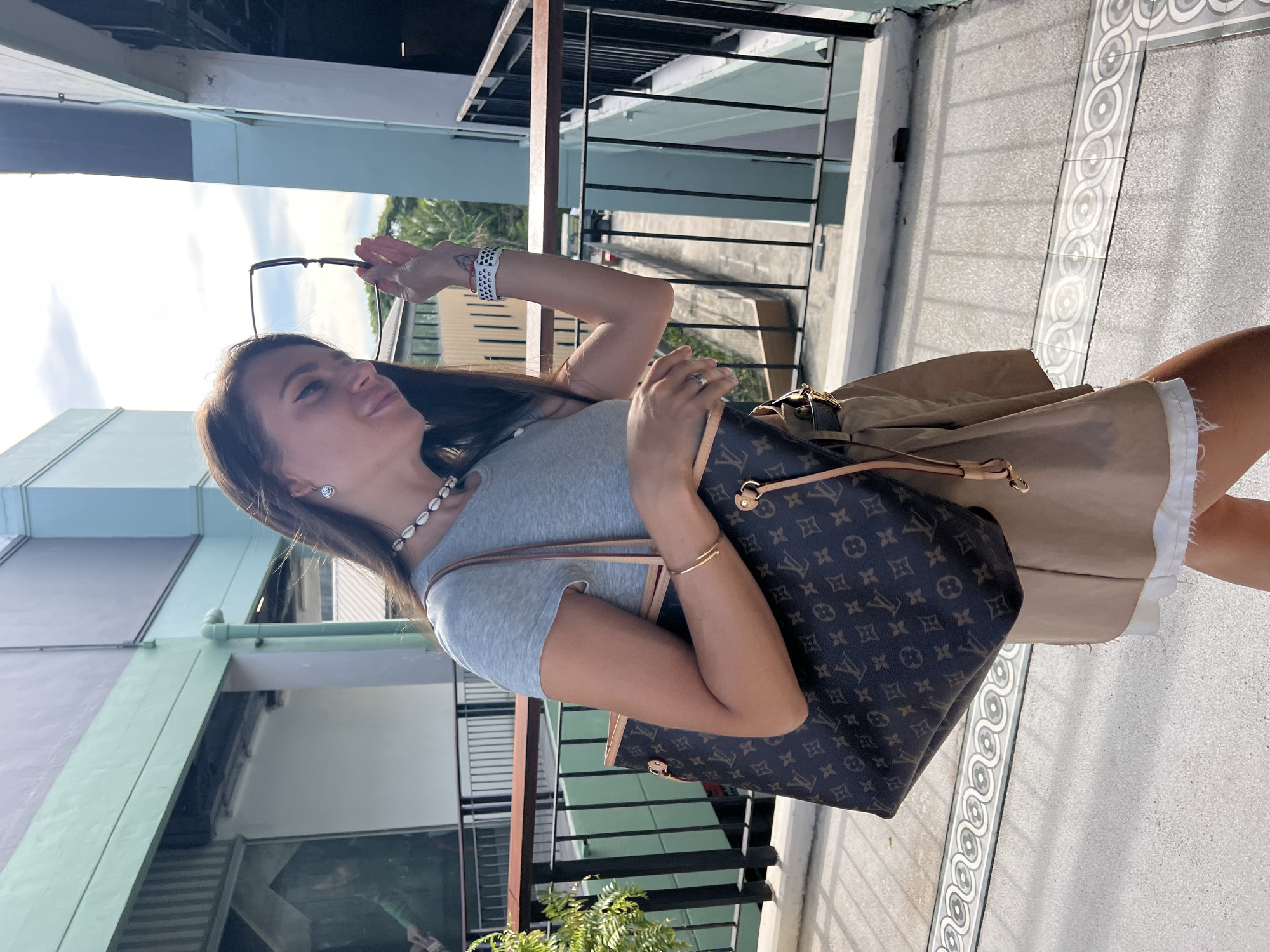 A woman walks along a balcony in Ban Bang Rak, Thailand, carrying a brown Louis Vuitton tote bag. She wears a grey t-shirt, a tan skirt over a white underskirt, a seashell necklace, a gold bracelet, and a smart watch. The woman holds a pair of glasses in her right hand, appearing to put them on. The environment includes a building with light blue and green accents, black railings, and a patterned tiled floor. Lush green foliage is visible in the background. The sky is overcast with bright sunlight.