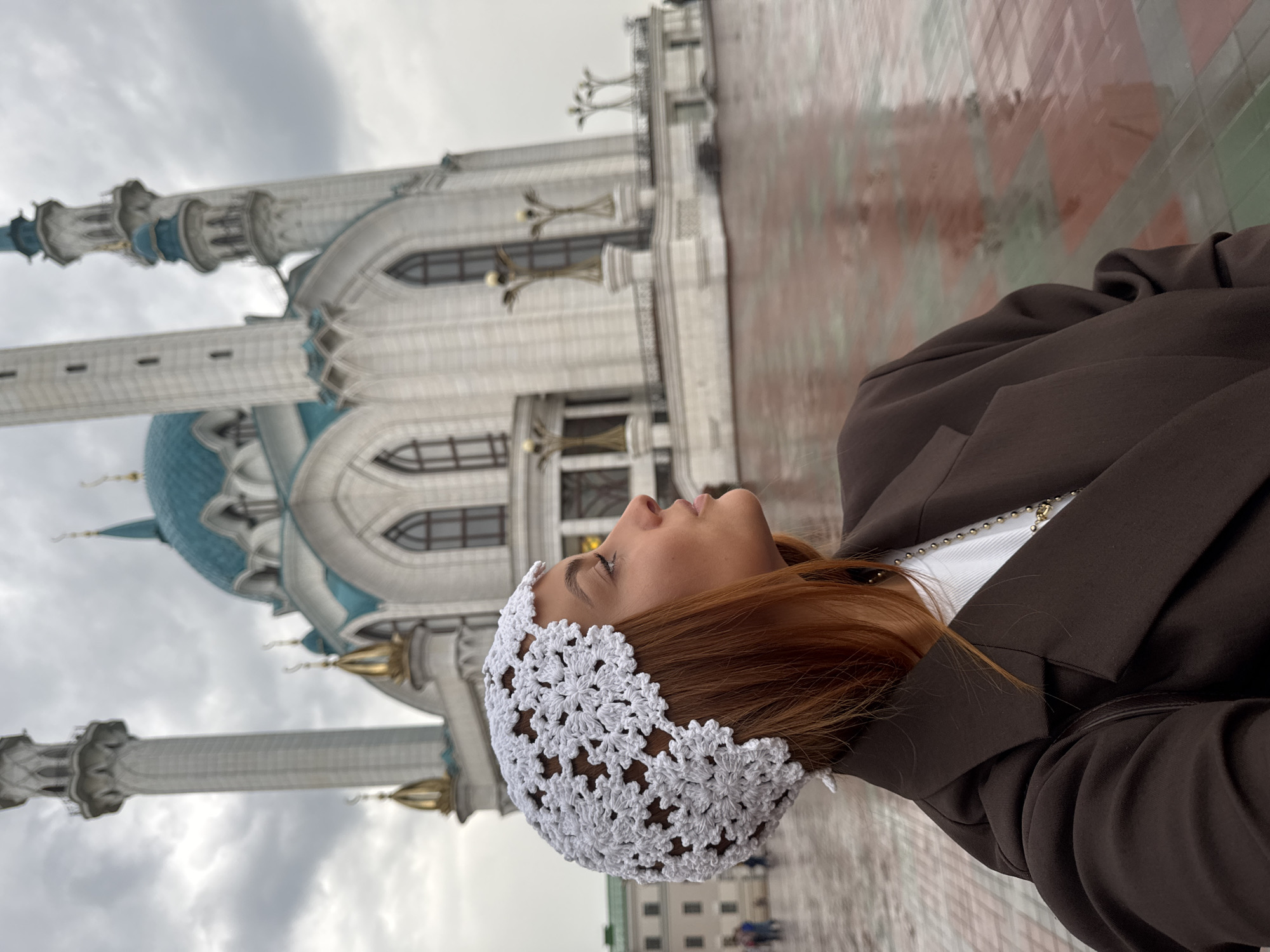 The image captures a woman with auburn hair, wearing a white crocheted headscarf and a dark brown blazer over a white top and necklace. She is looking to the right, with the Kul Sharif Mosque in Kazan, Russia, visible in the background. The mosque features white walls, a turquoise dome, and tall minarets. The ground in front of the mosque appears wet, suggesting recent rain, and is paved with red and green tiles. Several lampposts are visible on the mosque's elevated platform. In the far background, other buildings and a few people can be seen. The sky is overcast and cloudy.
