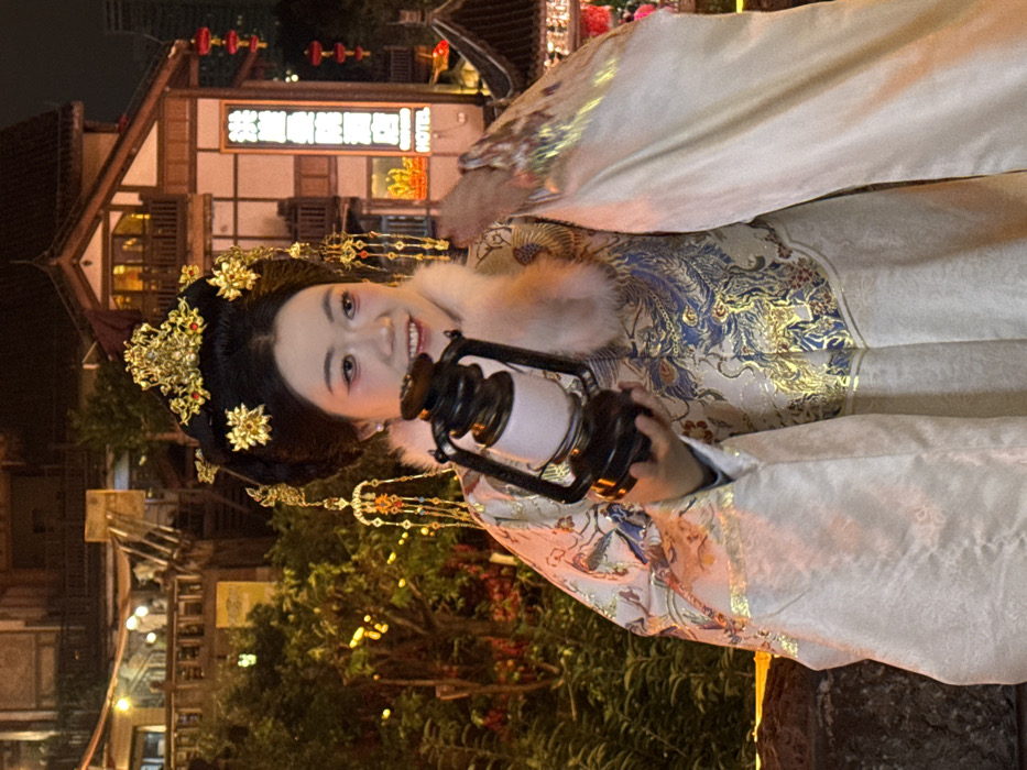 An adult female is depicted at night in Yuzhong District, China, positioned slightly right of the frame's center. She is smiling and looking towards the right. She wears elaborate traditional Chinese attire, featuring a cream-colored outer robe with significant gold and blue embroidered patterns, possibly depicting mythical creatures, over an undergarment. A light-colored, fur-trimmed collar is visible. Her hair is styled intricately with a prominent golden headpiece adorned with red accents, and additional golden floral hairpins and dangling ornaments with red beads. She holds a dark-toned lantern with a light-colored cylindrical shade in her right hand, held near her chest. The lantern appears illuminated. The background features multi-story traditional Chinese buildings with warm interior lighting and balconies. One building on the right has a vertical neon sign with Chinese characters and the word "HOTEL" displayed in English and Chinese. Red Chinese lanterns hang from this building. Dark foliage with numerous small, warm string lights is present behind her. A dark, textured surface, possibly a stone railing, is visible in the lower left foreground.