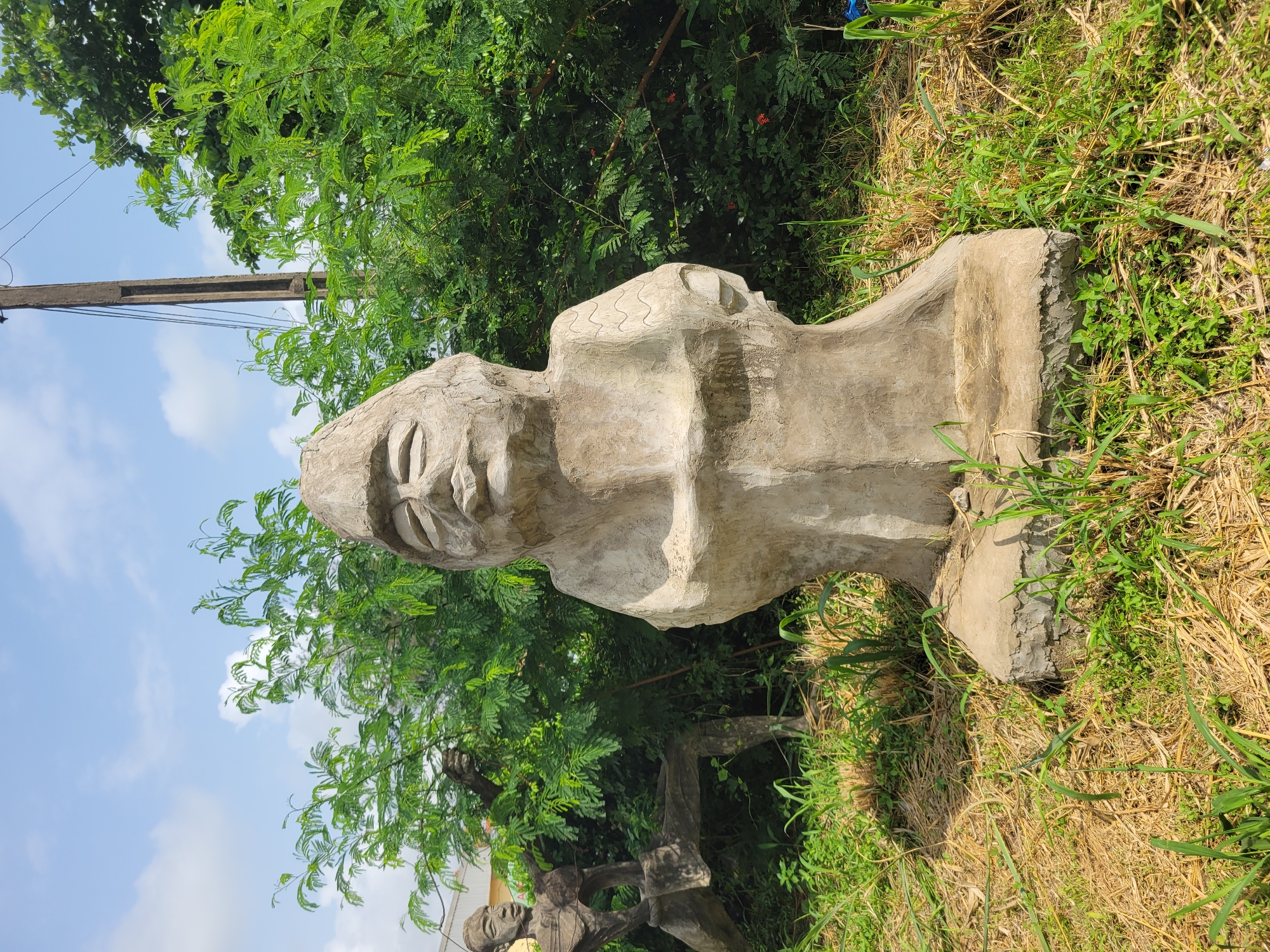 The image depicts an outdoor scene in Egbagu, Nigeria, featuring two stone sculptures. In the foreground, a larger sculpture of a seated figure with a distinct hat and facial features is the central focus. The figure is positioned on a concrete base amidst dry grass and greenery. Behind this sculpture, to the left, a second, more abstract sculpture of a human-like form in a dynamic pose is partially visible, leaning against a tree. The background is filled with lush green foliage, a utility pole with wires, and a bright blue sky with scattered clouds. The overall setting suggests an artistic installation or a place of cultural significance in a natural environment.