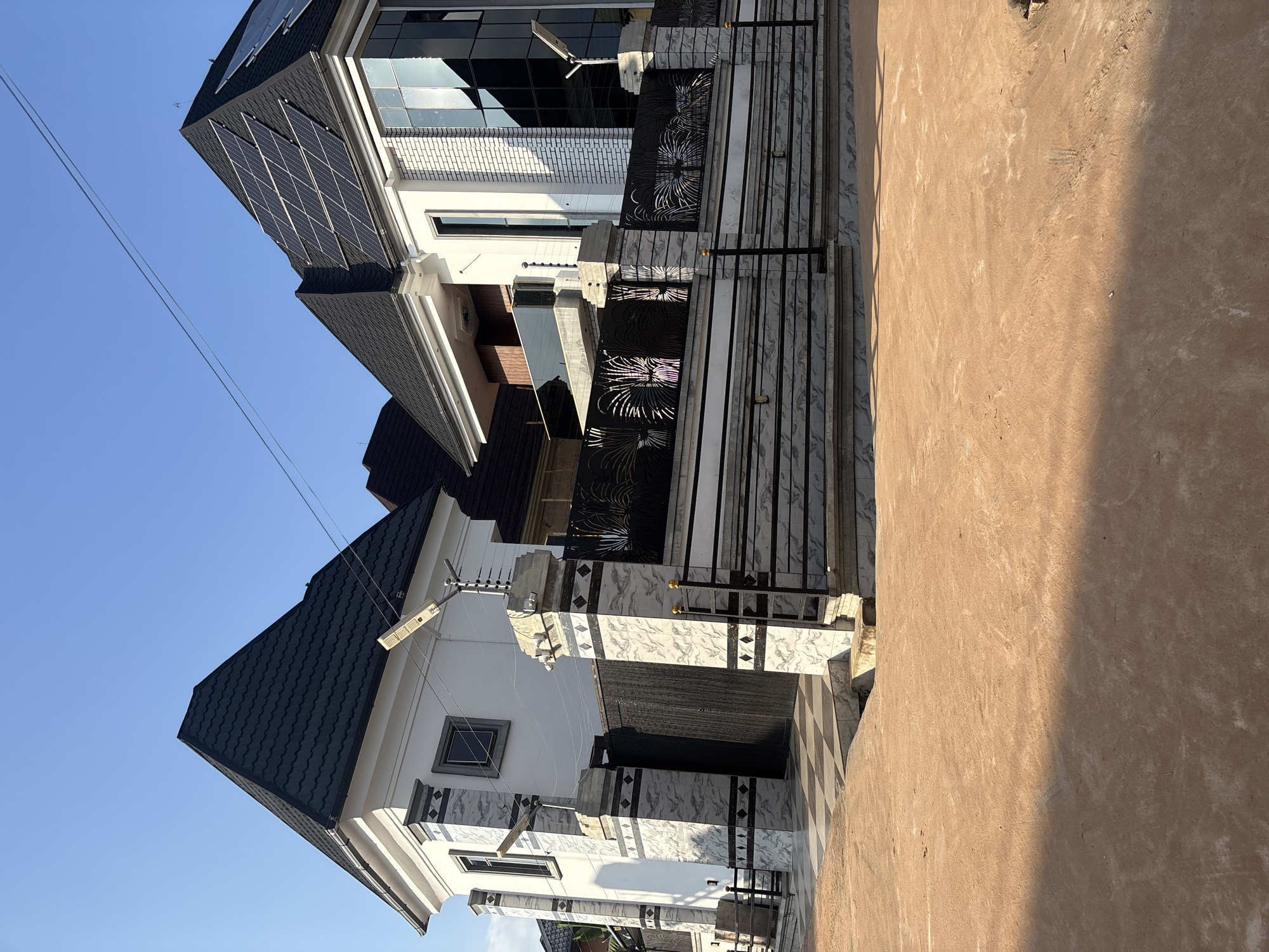 In Obosi, Nigeria, a modern duplex with a dark-tiled, steeply pitched roof stands under a clear blue sky. Solar panels are visible on the roof of the right side of the duplex, which features a white brick facade and large, reflective windows. The duplex is bordered by a decorative black metal fence with an intricate pattern, supported by pillars clad in grey and white marbled tiles. A tall, dark metal gate is partially visible behind the fence. The ground in front of the duplex is a mix of reddish-brown earth and a checkered tiled area leading to the gate. The scene appears to be on a sunny day, with long shadows cast across the ground.