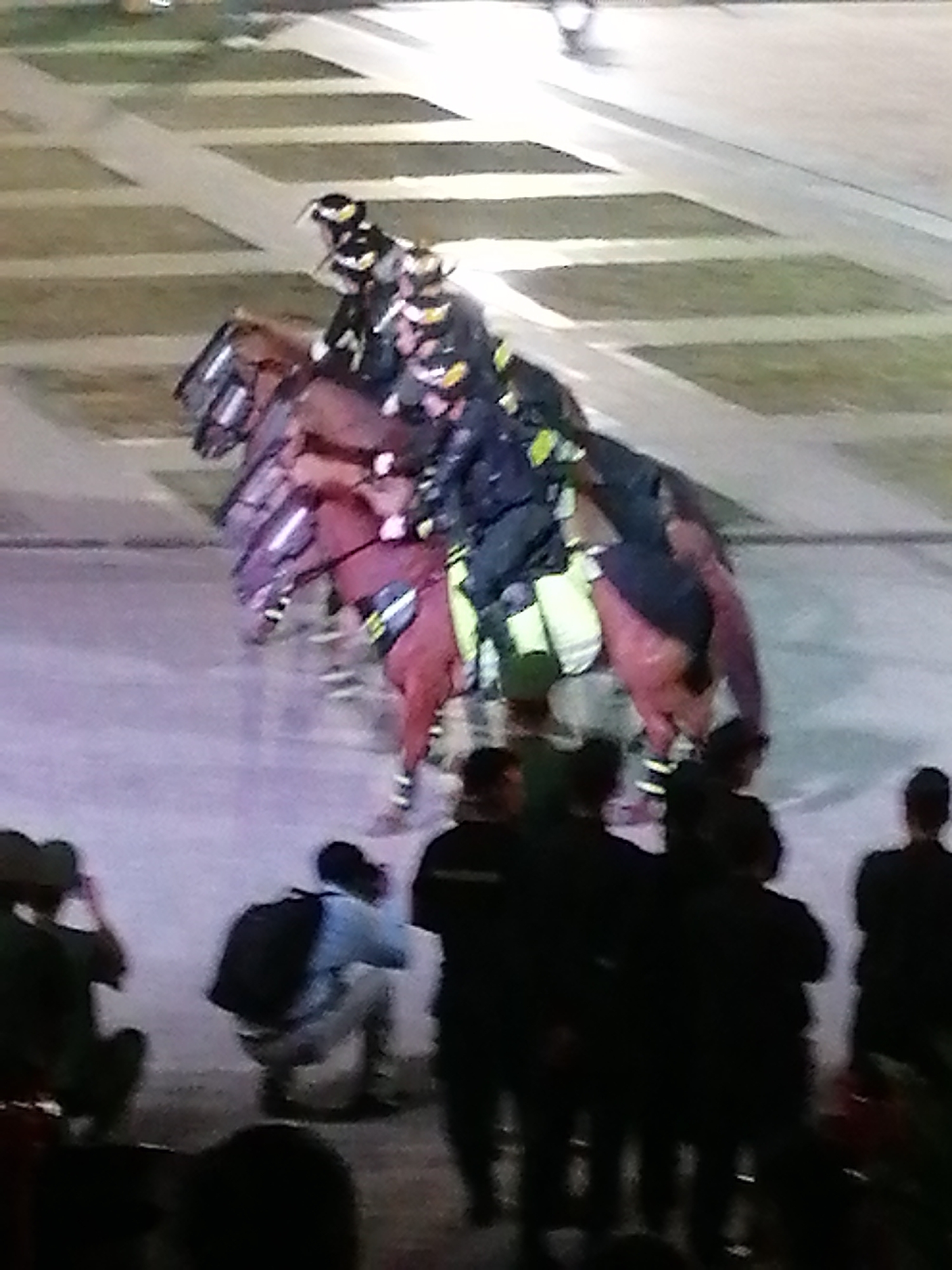 The image depicts a group of individuals on horseback, seemingly participating in an organized event or parade. The riders are dressed in what appear to be uniforms, with helmets and distinctive clothing, suggesting they might be part of a police or ceremonial unit. The horses are adorned with what look like protective gear or decorations. In the foreground, a crowd of people is visible, some seated and others standing, observing the activity. One individual in the crowd is crouched down, wearing a backpack. The setting appears to be an outdoor venue with paved surfaces and some grassy areas, possibly a park or a public square. The lighting suggests it is either dusk or a dimly lit evening. The overall atmosphere seems calm, with the focus on the equestrian display. No visible text is present in the image. The location context of Da Lat, Vietnam, might suggest a cultural event or a formal occasion.