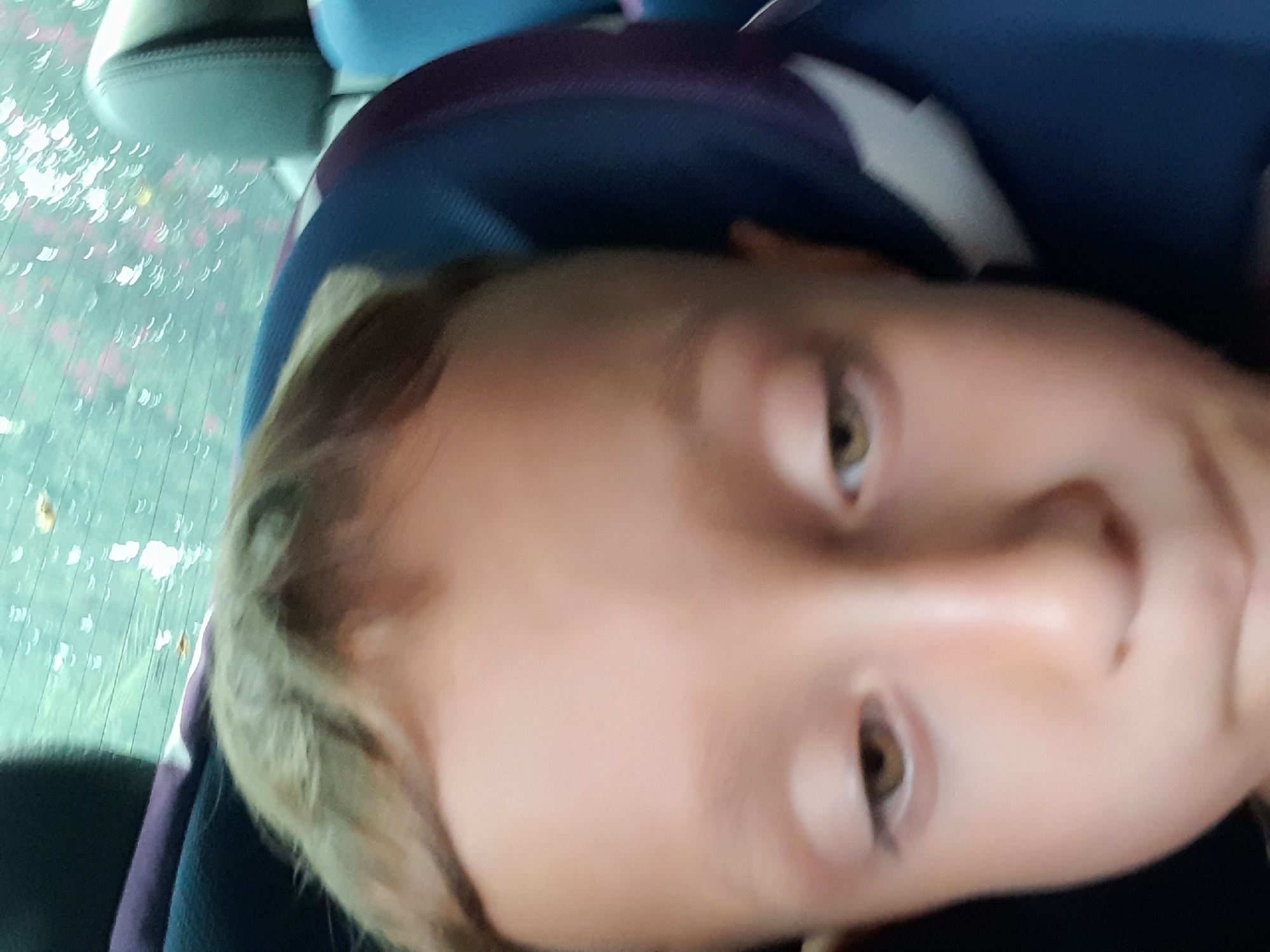 The image is a close-up, low-angle, blurred selfie of a child's face. The child has light brown hair, fair skin, and brown eyes. They appear to be in a vehicle, possibly a car or van, with a headrest and part of a seat visible. The background shows a window with water droplets and some out-of-focus foliage, suggesting it might be raining or the car is moving. The scene is indoors. There is no discernible city or location information present in the image itself.