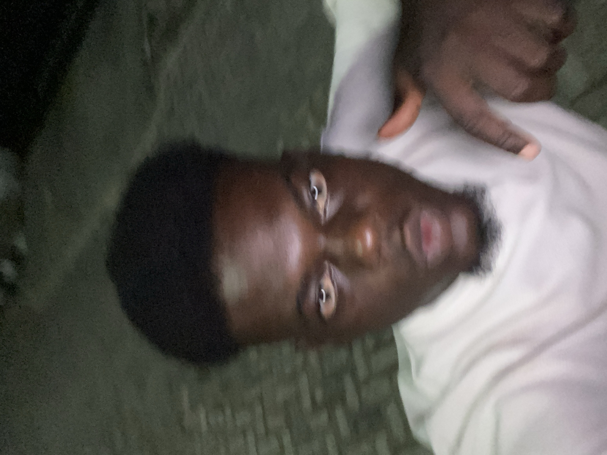 A selfie of a Black man wearing a white shirt is taken from a low angle. The man has short black hair, dark eyes, and a short beard. His expression is inquisitive. A hand, likely belonging to the man taking the photo, is visible in the upper right corner. The background is indistinct but appears to be a textured surface, possibly a pavement. The location is unknown.
