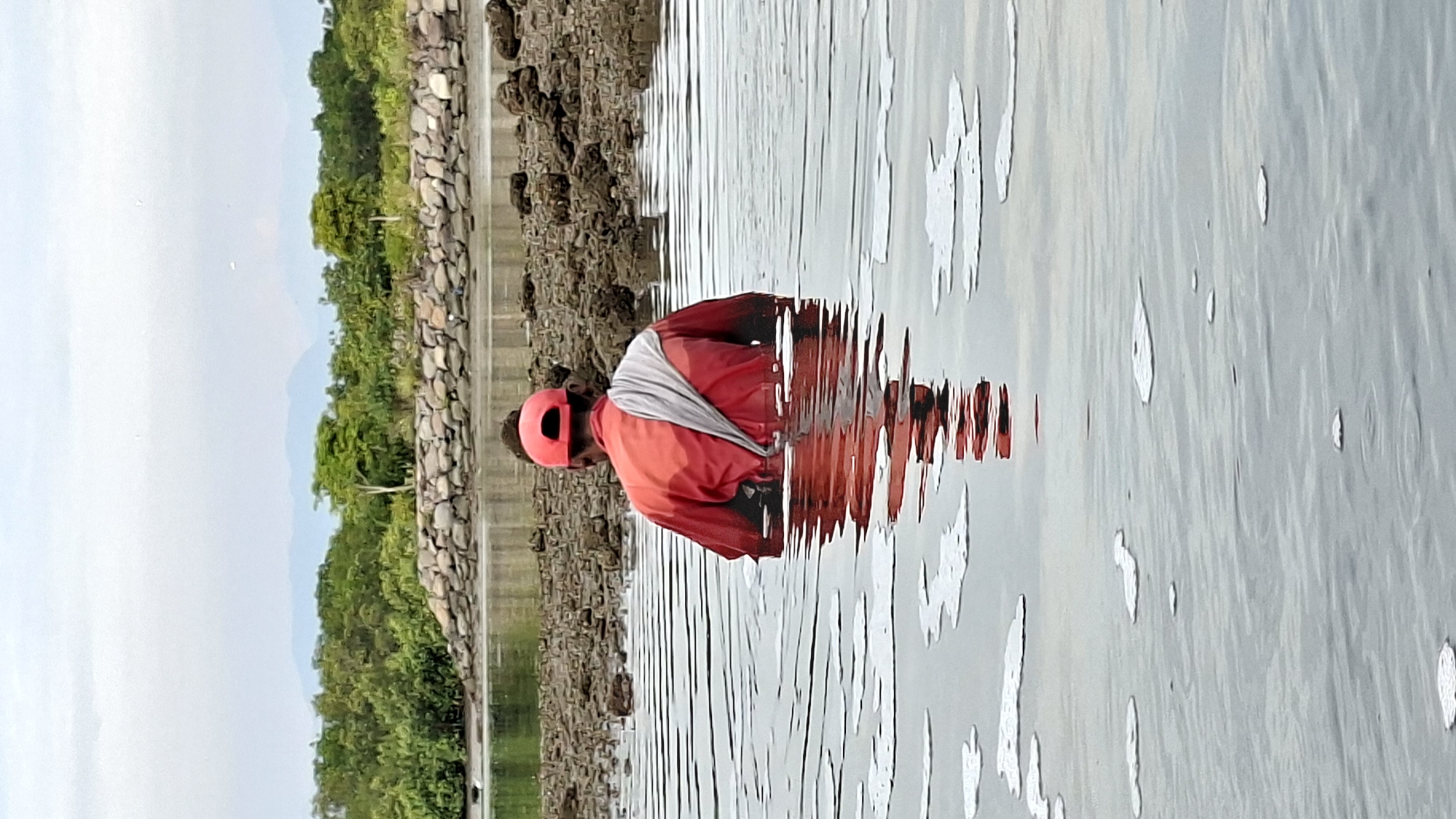 A person wearing a red jacket and a red helmet is standing in shallow water. The person is facing away from the camera, and their reflection is visible in the water. The background consists of a rocky shoreline with lush green trees and vegetation. Mountains are visible in the distance under a bright, overcast sky, suggesting it is daytime. The water is calm, with ripples distorting the reflection of the person. The scene appears to be a coastal area in Lhokseumawe, Indonesia, possibly a beach or a shallow bay. There is no visible text in the image.
