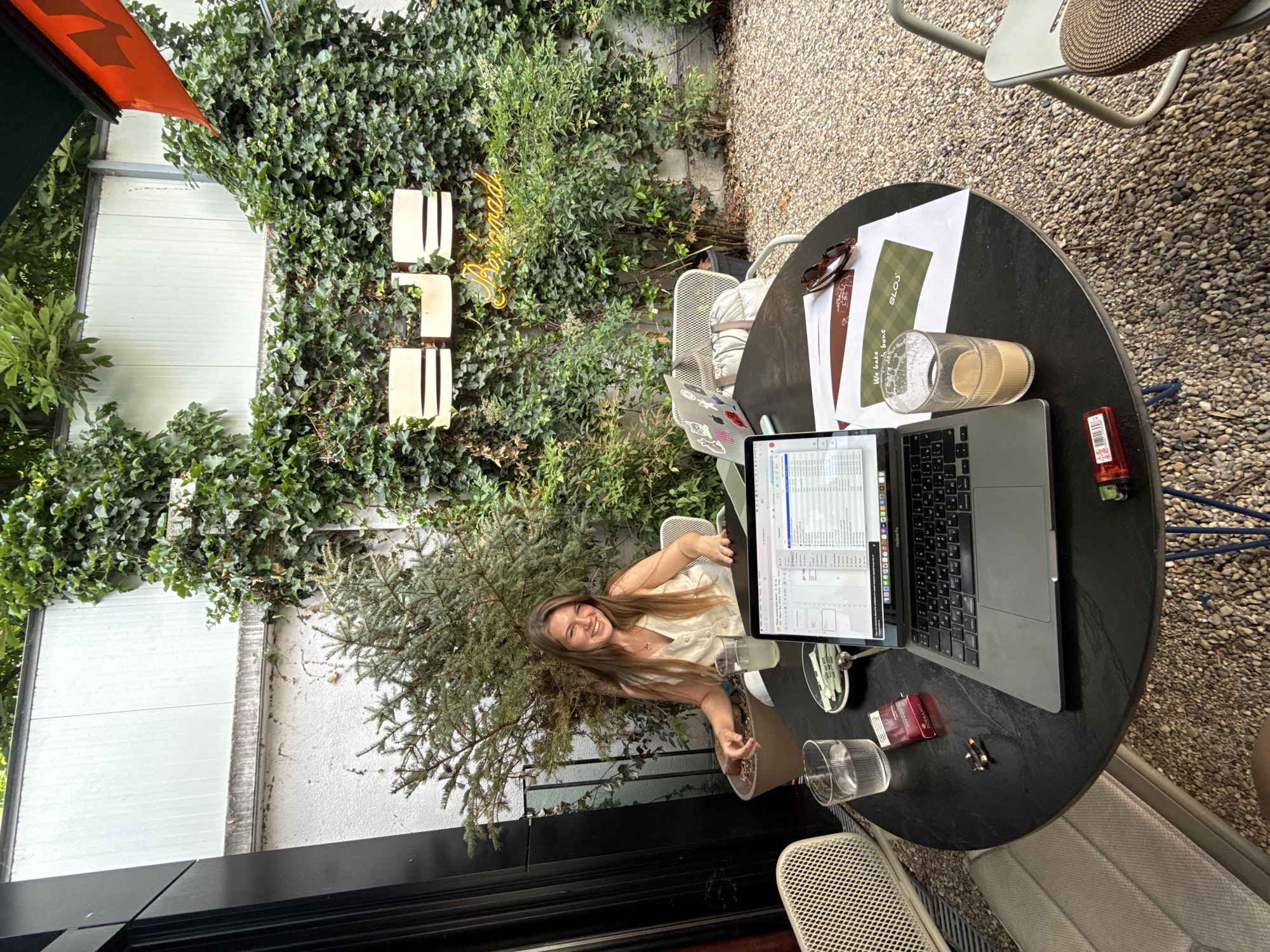 A young woman with long, light-brown hair is sitting at a black, round table in an outdoor setting, possibly a café or restaurant. The table is laden with items including a laptop, documents, a glass of liquid (possibly coffee), a lighter, and cigarette packs. The woman is smiling and looking at the camera. The environment features dense greenery, including ivy-covered walls and small trees. The ground is covered with gravel.
Based on the documents on the table which reference "We bake very Sloj..." and the "SLOJ" logo, this is likely a café in Belgrade, Serbia called "SLOJ". Additionally, there is a sign that says "homemade" in the ivy-covered wall on the background.