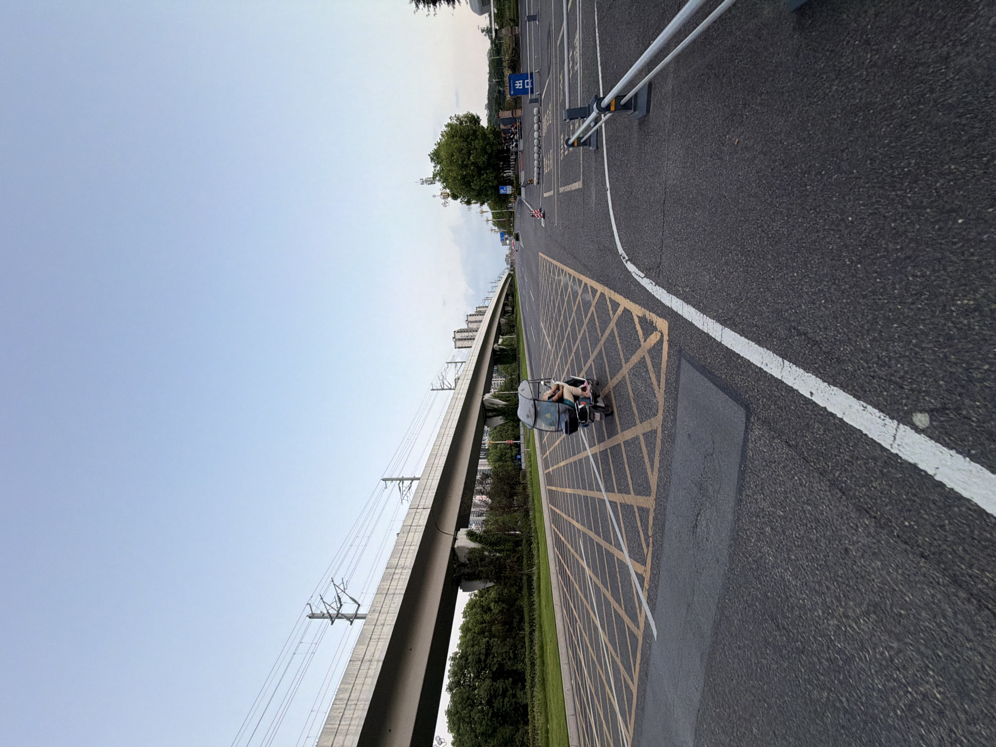 An outdoor, wide-angle, eye-level shot captures a road with a person riding a three-wheeled electric scooter. The scooter, a dark color with a canopy, has a single occupant. The road is paved asphalt and is marked with yellow crosshatching in a diamond pattern, indicating a no-parking zone. A white line demarcates the edge of the lane. In the background, an elevated concrete structure, likely a bridge or overpass for a railway or highway, runs horizontally across the image. Support pillars for this structure are visible. Beyond the structure, a line of lush green trees and residential buildings can be seen. The sky is clear with a pale blue hue, suggesting it's either early morning or late evening. To the right of the road, there's a parking lot area with a barrier gate and a blue sign with Chinese characters that reads "出口" (chūkǒu), meaning "exit." Several spherical bollards are arranged in a row leading to the exit. A few motorcycles are parked in the background of the parking lot. The overall scene is a typical urban or suburban environment in Jiangjin, China, likely a public road or entrance to a facility.