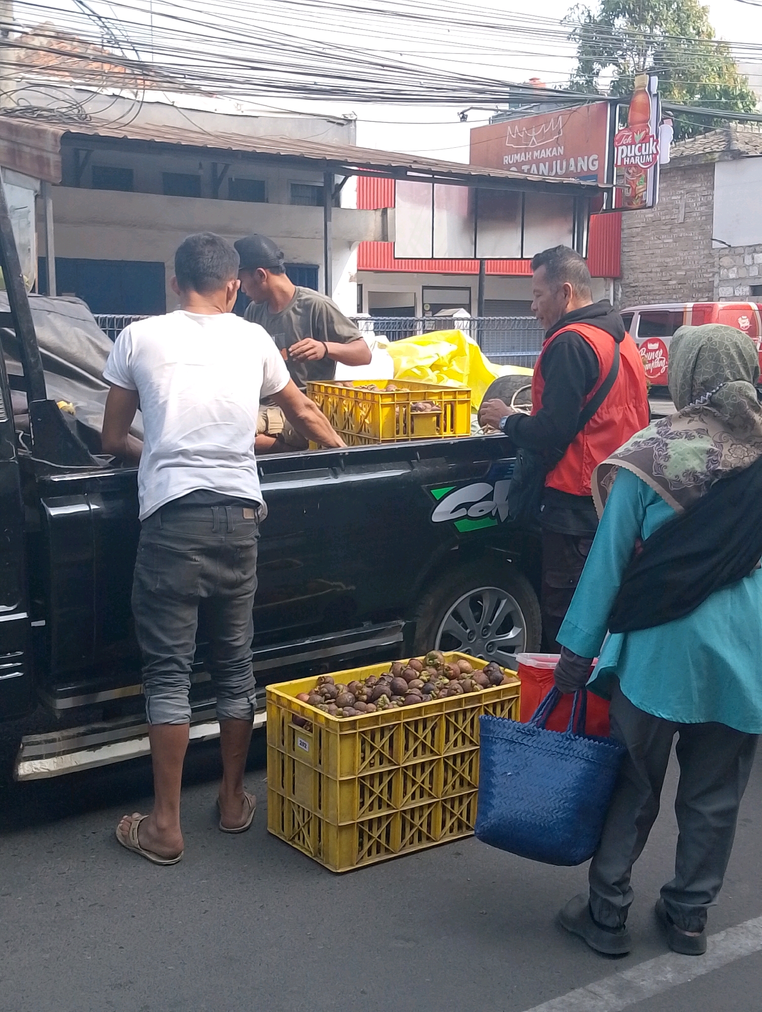 The image depicts a street scene in Lembang, Indonesia, with a focus on a fruit stand. In the foreground, a yellow crate overflowing with mangosteens sits on the asphalt. Behind it, a person wearing a teal shirt and grey pants carries a blue woven bag and a red container. Further back, a man in a black jacket and red vest is visible. To the left, two men are seen near a black truck. One man, wearing a white t-shirt and grey pants, appears to be unloading items from the truck. The other man, wearing a dark t-shirt and a black cap, is interacting with the first man. Above, power lines crisscross the sky, and a sign for "Rumah Makan Tanjuang" and "Teh Pucuk Harum" is prominently displayed. The overall environment suggests a bustling commercial area with food vendors.
