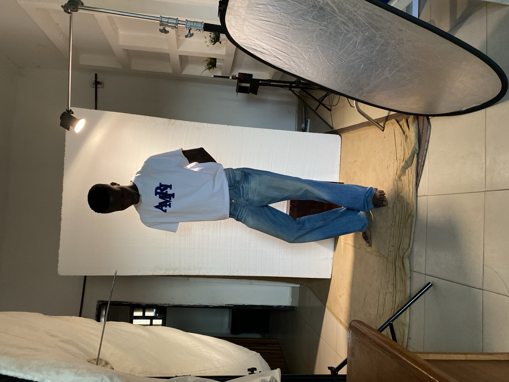 An indoor photograph captures a male subject posing in what appears to be a makeshift studio environment. The man, dark-skinned with a short haircut, stands centrally, facing slightly left, against a large white backdrop. He is wearing a white short-sleeved t-shirt with the word "AMIRI" printed in blue capital letters on the chest, paired with light blue jeans. He is barefoot.
The setting features various photography equipment. A spotlight mounted on a boom arm illuminates the subject from the left side of the frame, casting a bright light on the white backdrop. To the upper right, a large, oval silver reflector or softbox stands on a light stand. The floor is covered with light-colored tiles, over which a tan-colored rug or fabric is laid, extending from beneath the subject's feet to the right.
In the foreground, to the left, part of a bed with white bedding is visible. The background reveals architectural details of the room, including shelves with small decorative items, a section of a tiled wall, and a glimpse of a window with horizontal bars. A wooden railing or furniture piece is also visible in the bottom right corner. The scene depicts a planned photoshoot, indicated by the subject's pose and the arrangement of lighting equipment and backdrop.