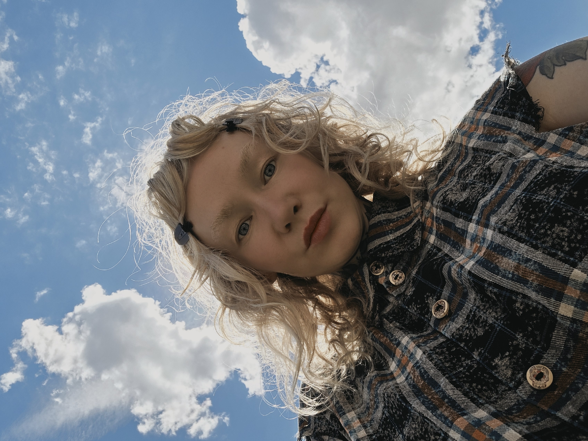 This is a selfie taken from a low angle, looking up at the subject against a bright blue sky with scattered white clouds. The subject is a young person with curly blonde hair, which is partially illuminated by the sun. They are wearing a plaid shirt with dark, blue, and orange tones. The shirt has several visible buttons, each with a skull design. A tattoo is partially visible on the subject's upper arm. The lighting suggests it is daytime, and the clear sky indicates pleasant weather. The camera is tilted, creating a dynamic perspective. The overall mood is casual and perhaps slightly playful due to the upward angle and the subject's direct gaze. No visible text is present in the image.