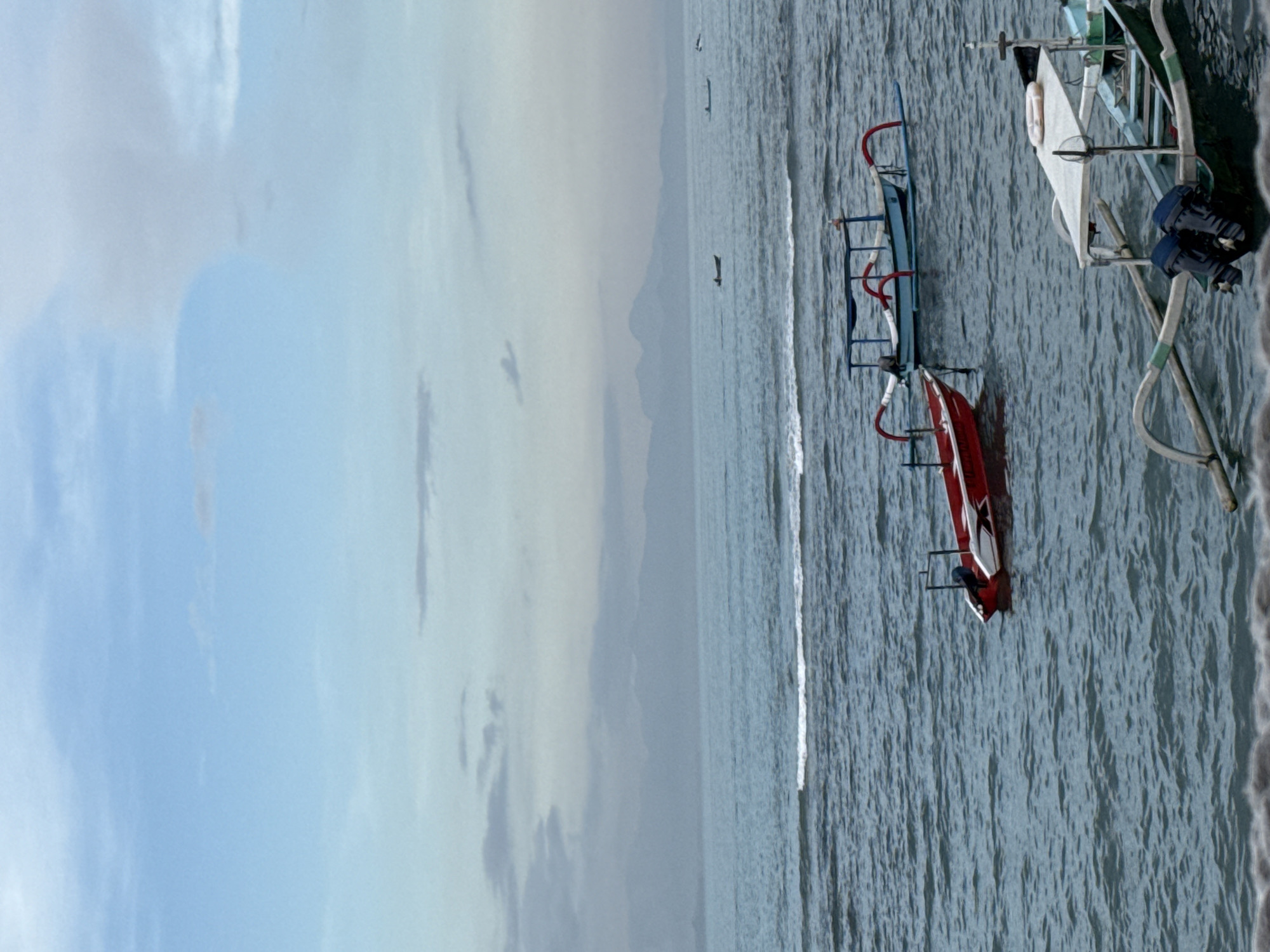 The image shows a seascape near Denpasar, Indonesia. The sky is overcast with a mix of blue and grey clouds. A beach is visible along the right edge of the frame, bordered by ocean water. Several boats are anchored in the water, including a blue and red outrigger canoe. Other watercraft are also present. Two figures can be seen in the water at a distance.