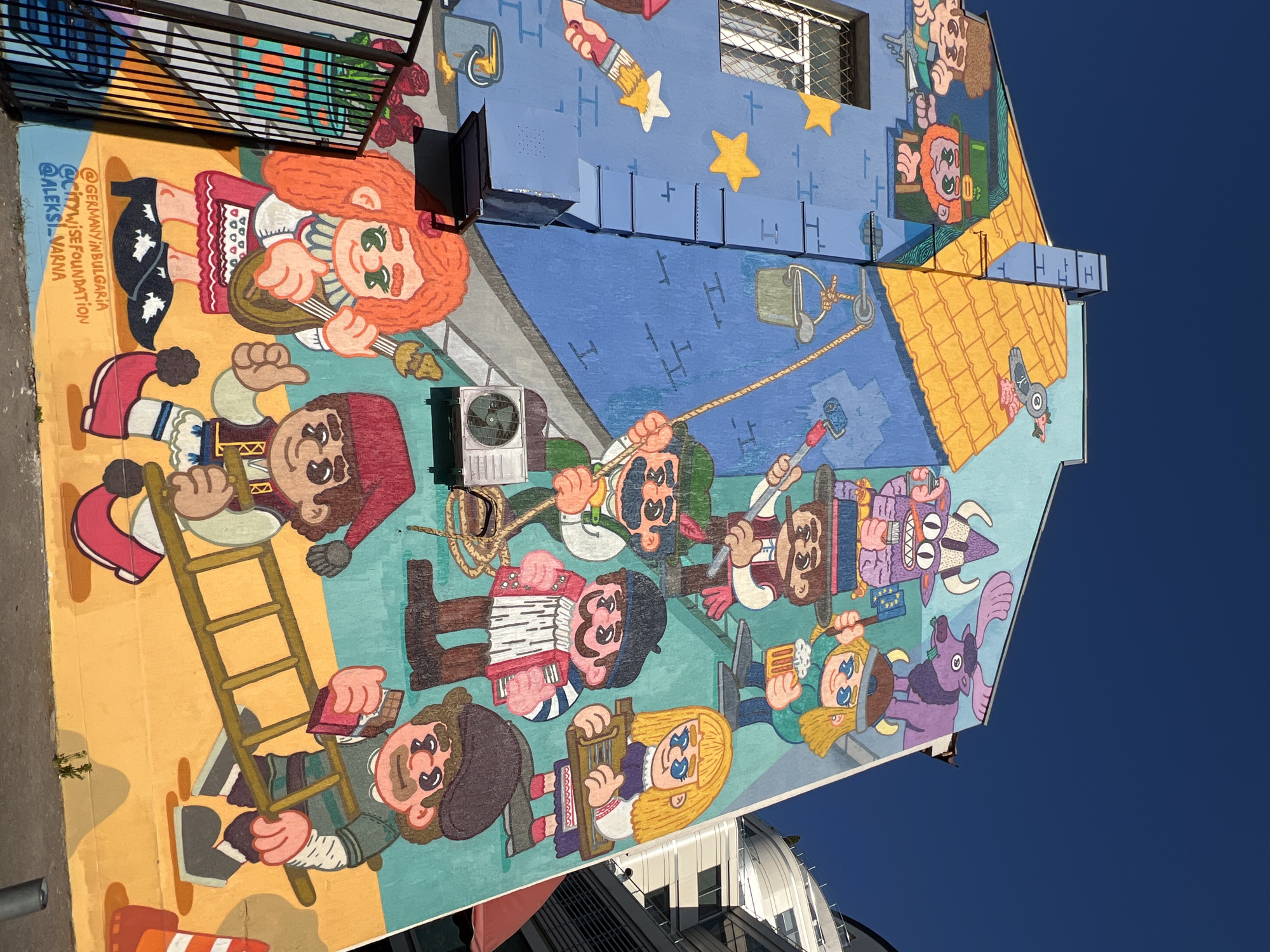 This is a photograph of a vibrant, colorful mural painted on the side of a building in Sofia, Bulgaria. The mural depicts a whimsical scene with cartoon-like characters. The characters appear to be engaged in various activities, such as holding musical instruments, tools, and food. There are also fantastical creatures and animals integrated into the artwork. The building itself has a blue facade with a yellow, tiled roof. The mural is detailed and covers a significant portion of the building's exterior. The sky above is a clear, deep blue. In the foreground, a black metal fence runs along the bottom of the image, partially obscuring the lower part of the mural. Text is visible at the bottom of the mural, indicating social media handles: "@GERMANYINBULGARIA", "@CITYWISEFOUNDATION", and "@ALEKSI_VARNA". These likely represent the artists or organizations involved in creating the mural.