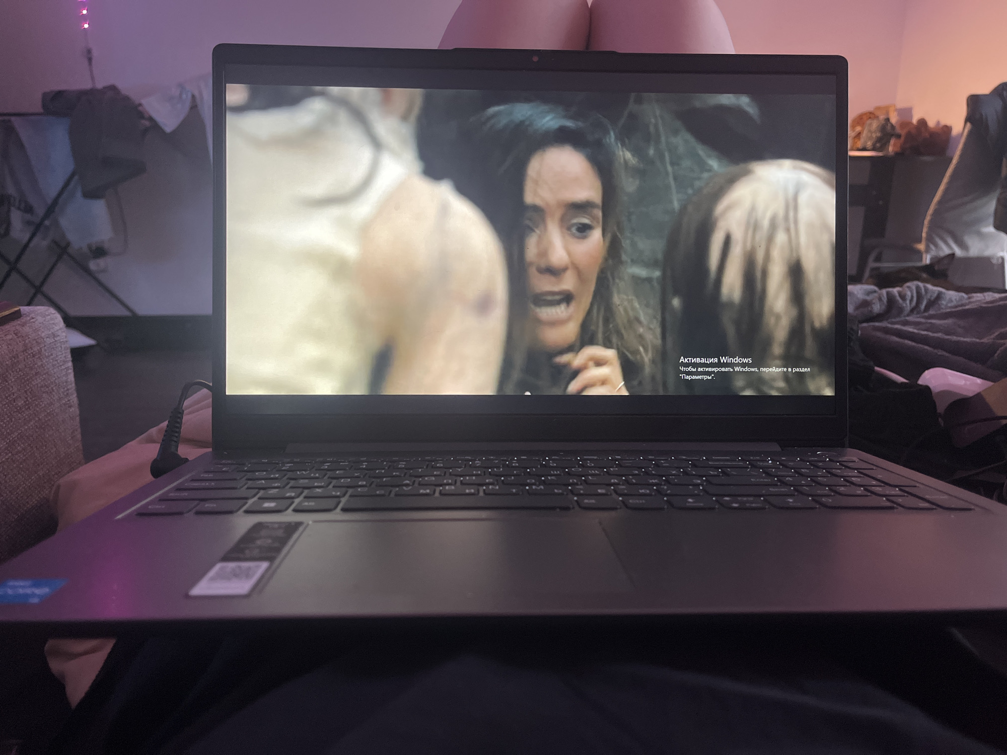 A laptop rests on someone's lap, displaying a scene from a movie or television show featuring a woman with a distressed expression. The laptop screen also shows a Windows activation notification in Russian. The background of the shot reveals a room with clothes, a bedside table, and various personal items. The location is indeterminate.
