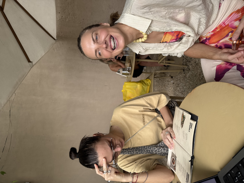 The image captures two adult females seated at a light brown round table within a cafe setting in Uluwatu, Indonesia. A third individual is partially visible in the background.
The woman on the left, with dark hair in a high bun, wears sunglasses, a light yellow "JACQUEMUS" t-shirt, multiple rings on her left hand, and bracelets on her left wrist. A leopard-print cross-body bag strap is visible. She is looking down at an open menu on the table, with her left hand touching her forehead. A silver laptop is partially visible on the table's bottom-left edge.
The woman on the right, with light brown hair pulled back, displays visible dental braces on her upper teeth as she smiles or laughs, looking towards the right out of frame. She wears a chunky light yellow beaded necklace and a light-colored, striped, unbuttoned long-sleeve shirt over a brightly patterned top or dress in shades of orange, pink, and white. She holds a transparent, possibly amber-tinted object in her right hand, which rests on her lap.
In the background, a partially obscured third individual is seated at a small table with a glass of water. A bright yellow bag rests on the floor next to this table. The background features a plain light beige wall and a white outdoor umbrella in the upper right corner, indicating a semi-outdoor cafe environment with natural lighting.