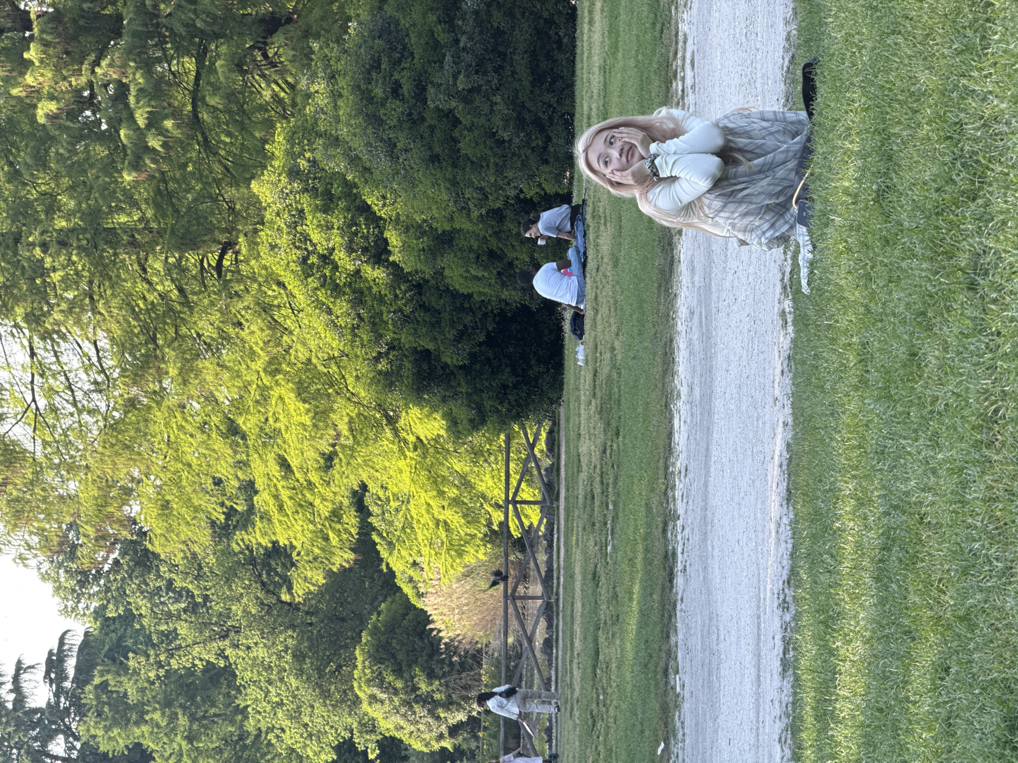 The image captures an outdoor scene in a vibrant green park, likely in Milan, Italy, on a sunny day. A wide, light-colored dirt or gravel path runs across the frame, dividing lush grassy areas.
In the foreground, to the right of the path, a young woman with long blonde hair is looking directly at the camera with a playful or surprised expression, her hands touching her cheeks. She is wearing a light-colored long-sleeved top and a grey and white plaid skirt. Her attire and cheerful demeanor suggest a relaxed outing.
Further back, on the grass to the left of the path, several other individuals are present. Two people are seen sitting or lounging, seemingly enjoying the weather; one wears a white t-shirt, and another is in a grey top. In the mid-ground, a rustic wooden fence stretches across the park, and a dark bird is perched on it. Another person is visible standing near this fence.
The background is dominated by an abundance of diverse green trees and dense foliage, indicating a well-maintained natural space. The bright sunlight illuminates the scene, suggesting it is late morning or afternoon. The overall atmosphere is peaceful and leisurely, with people enjoying the pleasant park environment. There is no visible text in the image.