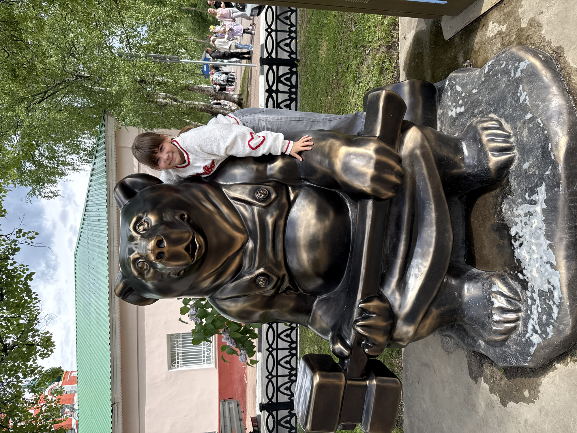 A bronze statue of a bear wearing a uniform, leaning on a hammer, stands on a concrete base outdoors. A girl in a white sweater and grey jeans leans on the back of the bear. In the background are trees, a building with a green roof, and a wrought-iron fence. A group of people is walking in the distance. The statue is located in Rusanovo, Russia.