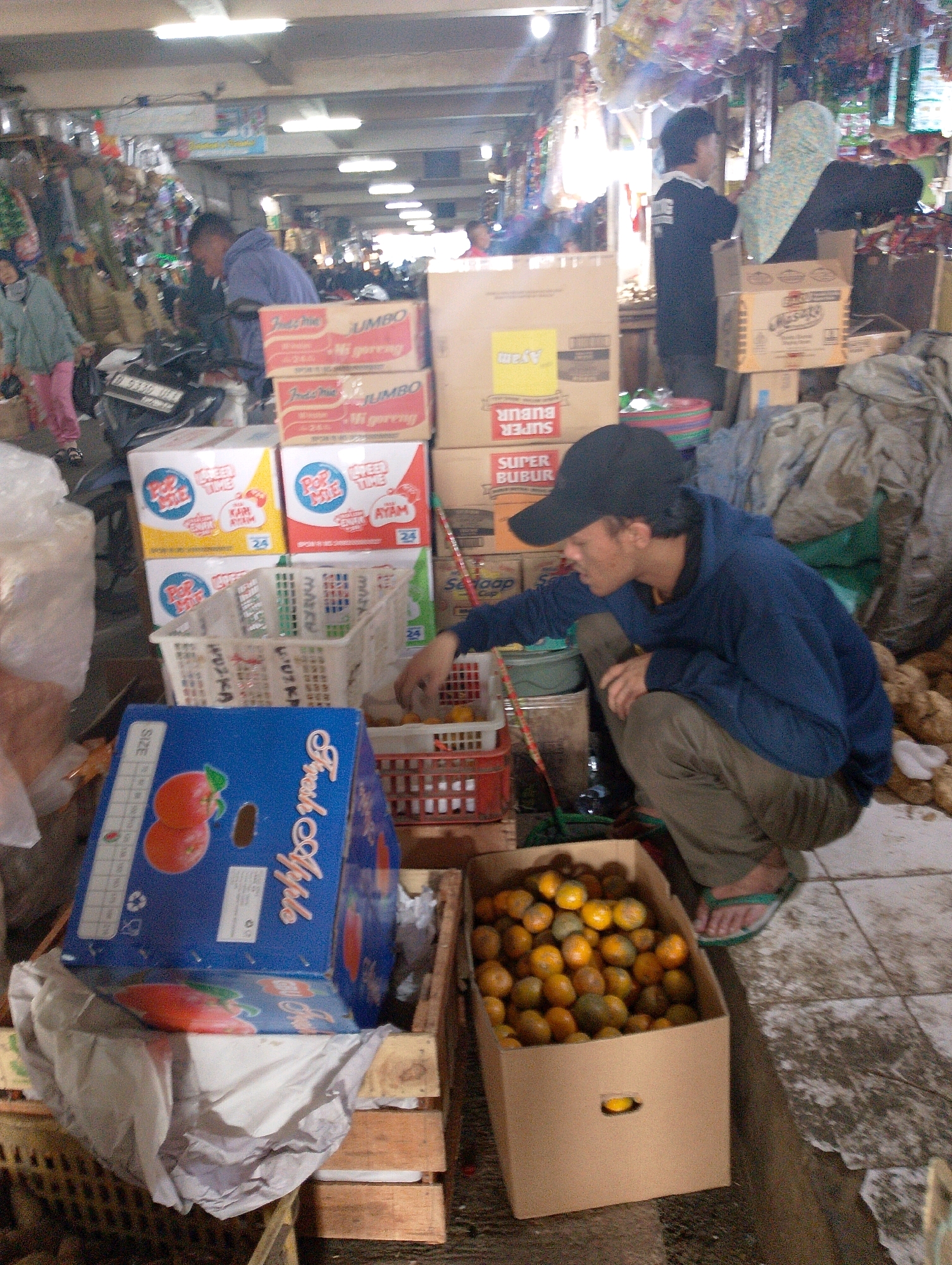 The image depicts a bustling market scene in Lembang, Indonesia. A young man wearing a blue hoodie and a black baseball cap is squatting down, looking at a box of oranges. He is wearing khaki pants and green flip-flops. Next to him, a large cardboard box overflows with oranges. In the background, stacks of packaged goods, including boxes of "Indomie" noodles and "Pop Mie" instant noodles, are visible. A woman in a light blue and white headscarf is standing behind the man, partially obscured. Other market stalls and shoppers are dimly visible in the distance. The ground appears to be tiled and slightly dirty. The overall impression is of a busy, open-air market environment.