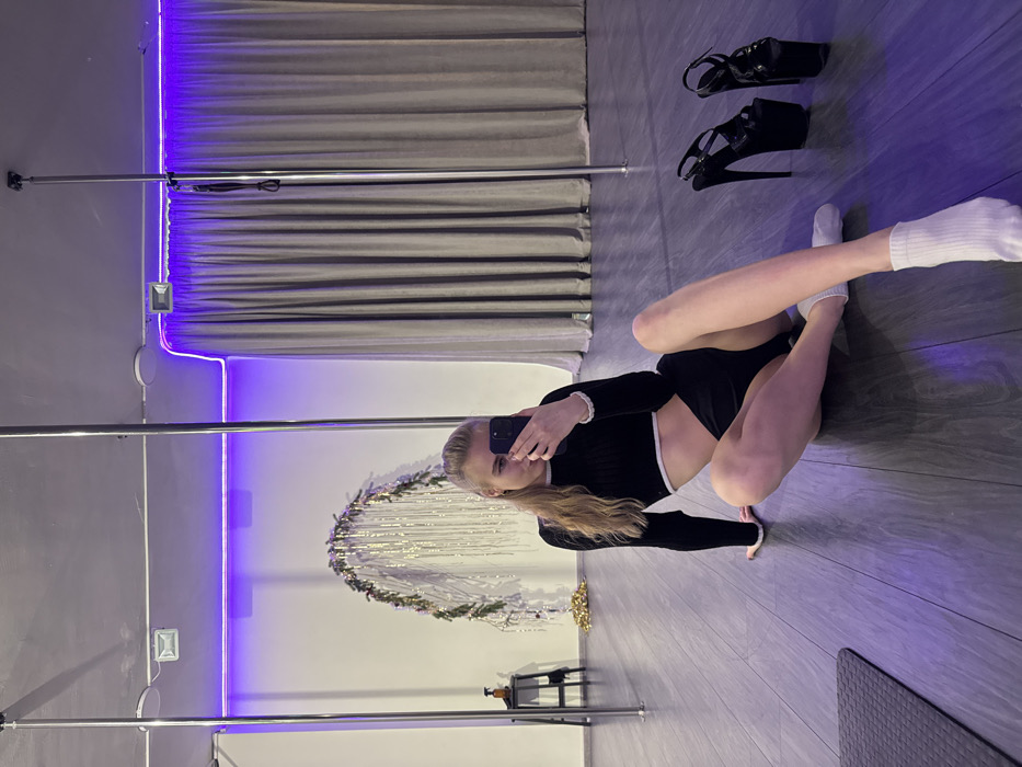 A woman in a black and white outfit and white socks poses in a dance studio, taking a selfie with her phone. Two tall metal poles stand behind her, and a gray curtain and glowing purple lights line the wall. A decorative circle with lights hangs on the wall as well. High-heeled black shoes are on the floor. The floor is wood-like and gray.