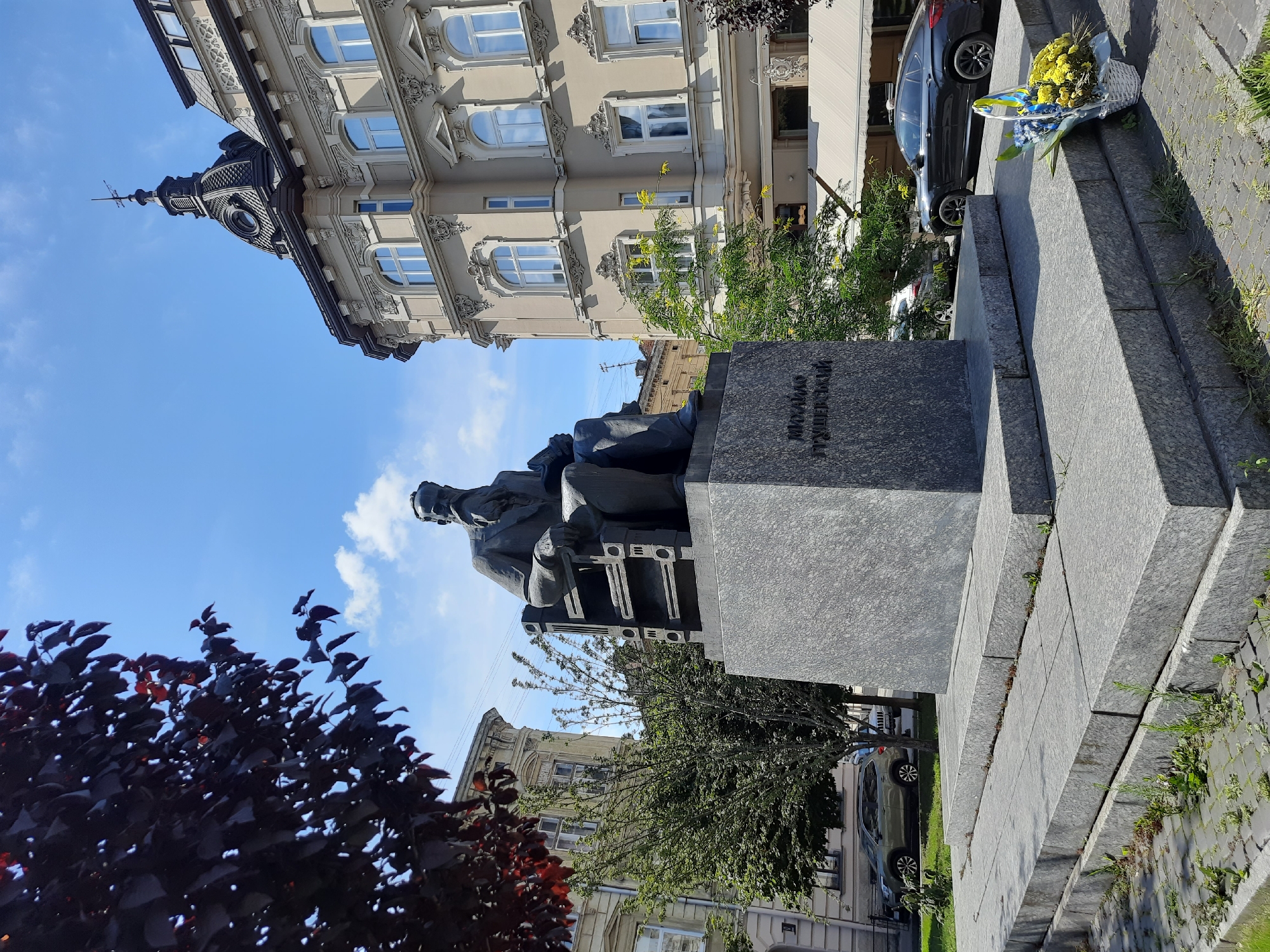 In Lviv, Ukraine, a statue of Mykhailo Hrushevsky, a prominent Ukrainian historian and politician, is situated in a public square. The bronze statue depicts Hrushevsky seated in a chair, with his signature beard and contemplative expression. The statue stands on a large granite pedestal, inscribed with his name. In front of the pedestal, a basket of flowers, adorned with a Ukrainian flag, rests on the steps, indicating a recent commemoration.
The background features an ornate architectural building with a distinct dome and spire, characteristic of Lviv's European-style cityscape. The sky is clear blue with a few scattered clouds, and trees with dark leaves frame the left side of the scene. Several cars are parked on the street behind the monument, and a light-colored awning extends from the building on the right, suggesting a commercial establishment. The ground is paved with cobblestones and stone slabs, with some grass and weeds growing through the cracks. The overall ambiance is one of historical significance and remembrance, set against a backdrop of urban beauty.