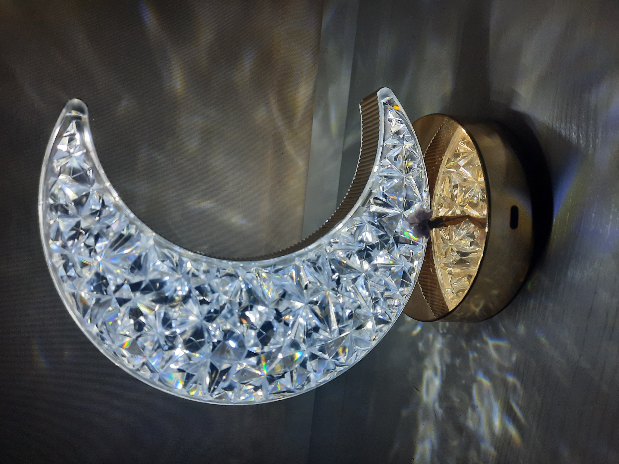 A crescent moon-shaped lamp with a faceted, crystal-like surface sits on a gold-colored base. The lamp is illuminated, casting colorful light refractions on the dark, textured surface it rests upon and the background. The background is dark and slightly textured, with faint, blurry rainbow-colored light patterns. The image does not provide any information about the city of Парэчча, Belarus.