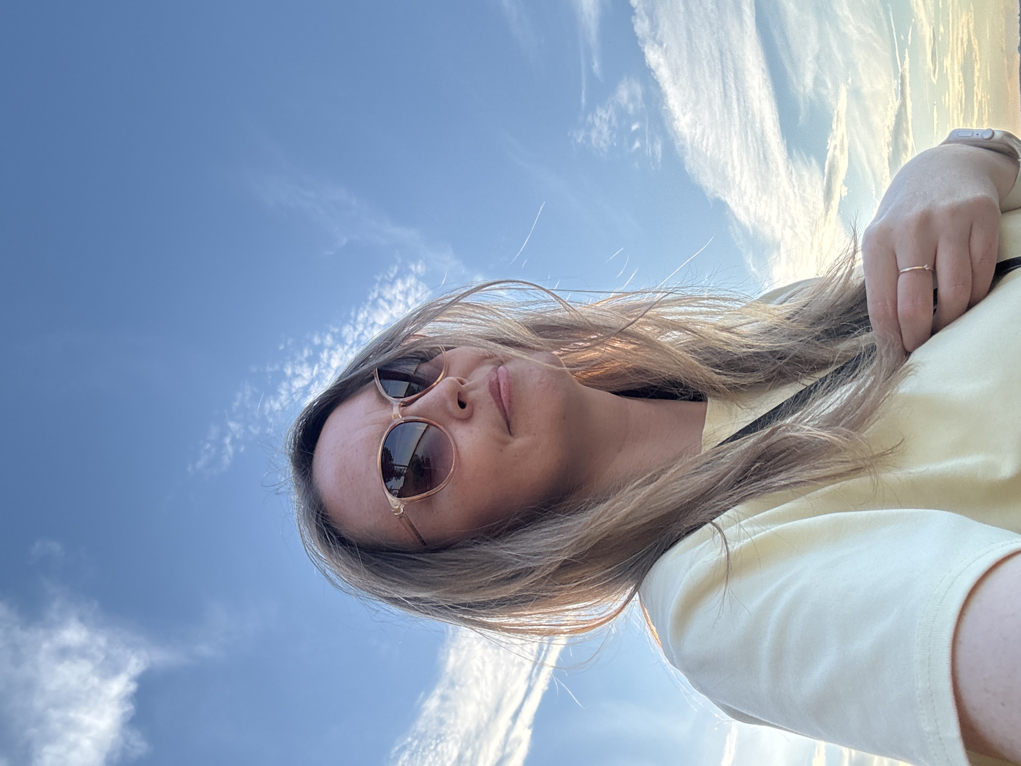 This media file is a low-angle, vertically oriented selfie of a young woman taken outdoors. She is wearing sunglasses and a light yellow t-shirt. Her hair is long and blonde, and it appears to be blown by the wind. Her left hand, adorned with a delicate ring, is visible in the upper right portion of the frame, resting on her clothing.
The background is a bright blue sky with wispy white clouds, suggesting a clear and sunny day. The lighting indicates it is likely daytime, possibly late afternoon or early evening, given the warm tones in the sky. There are no explicit location cues within the image itself that would pinpoint it to Chaikovsky, Russia, beyond what is provided in the prompt.
The woman's expression is relaxed and perhaps joyful, as if enjoying the outdoors. The angle of the shot and the wind in her hair contribute to a sense of freedom and spontaneity. There is no visible text in the image. The overall impression is of a pleasant, casual moment captured on a beautiful day.