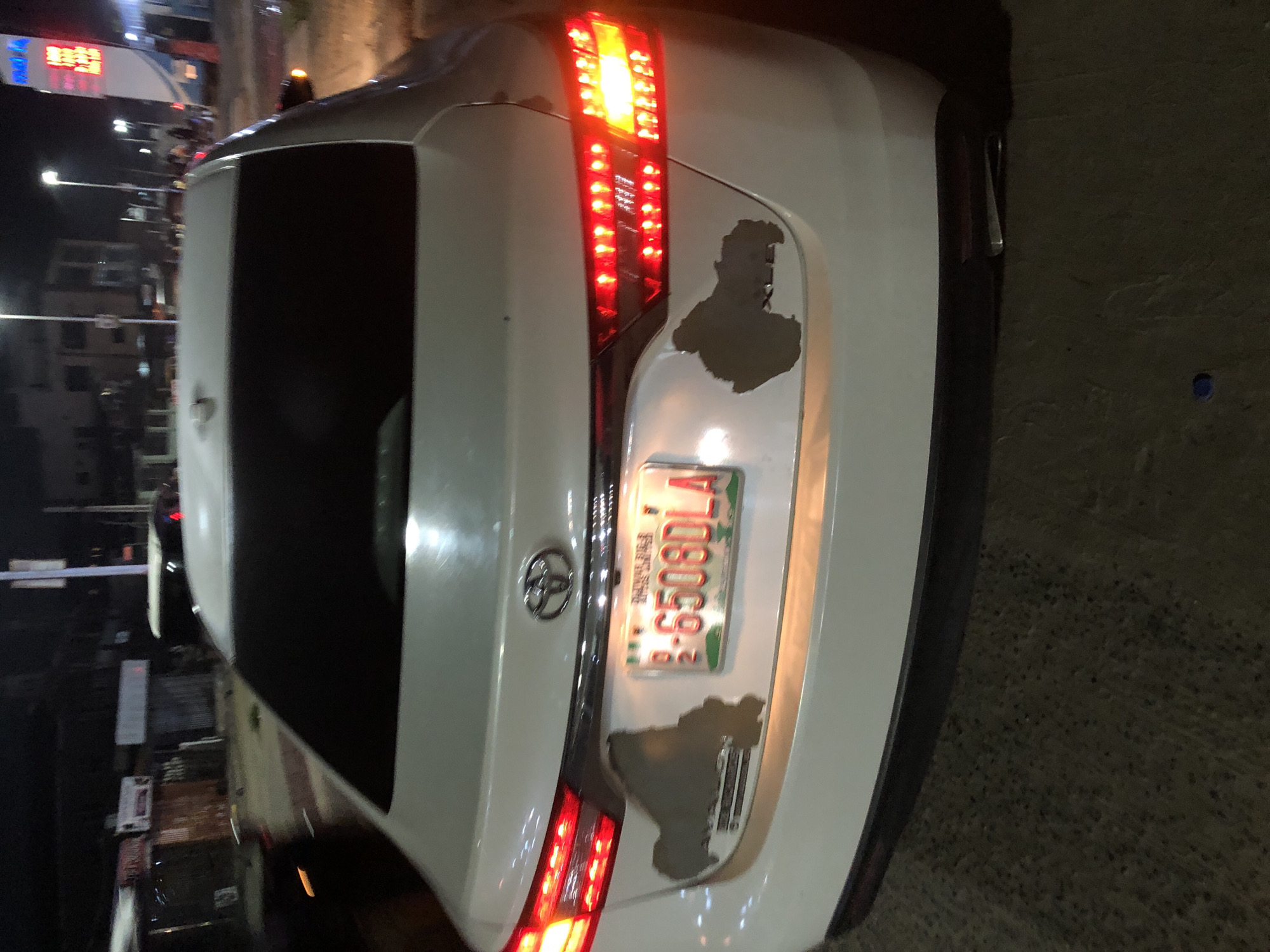 The image shows the rear of a white car with damage to the paint around the license plate. The license plate reads "R-6508DLA". The Toyota logo is visible above the license plate. The taillights are illuminated. The photo appears to have been taken at night in Ado, Nigeria, as indicated by the presence of a car with a Nigeria registration plate. The environment is dark with some streetlights and building lights visible in the background. The car is parked on a paved surface.