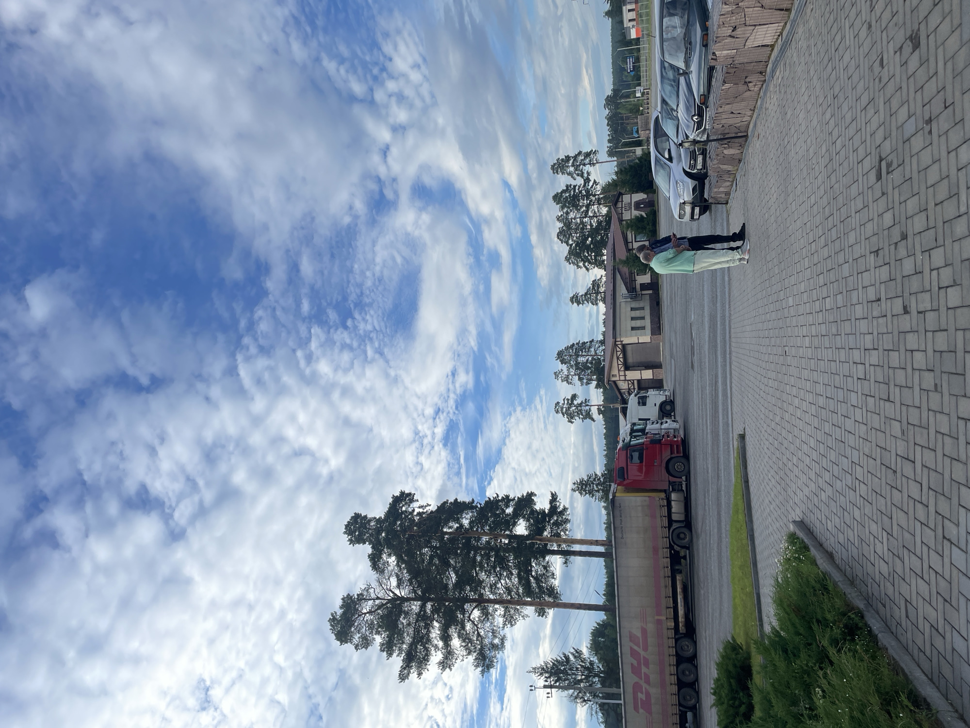 The media file shows a cloudy sky with scattered white clouds and a blue sky. In the foreground, there is a paved area with several cars parked in a row. To the right of the parked cars, a person is standing. Further back, there are buildings and trees. A red and white truck with a DHL logo on its trailer is parked on the paved area. The scene appears to be a parking lot or an area adjacent to buildings, likely in a semi-rural or industrial setting. The lighting suggests it is daytime, possibly late afternoon or early evening. The weather is cloudy but appears to be fair. The DHL logo is visible on the side of the truck trailer.