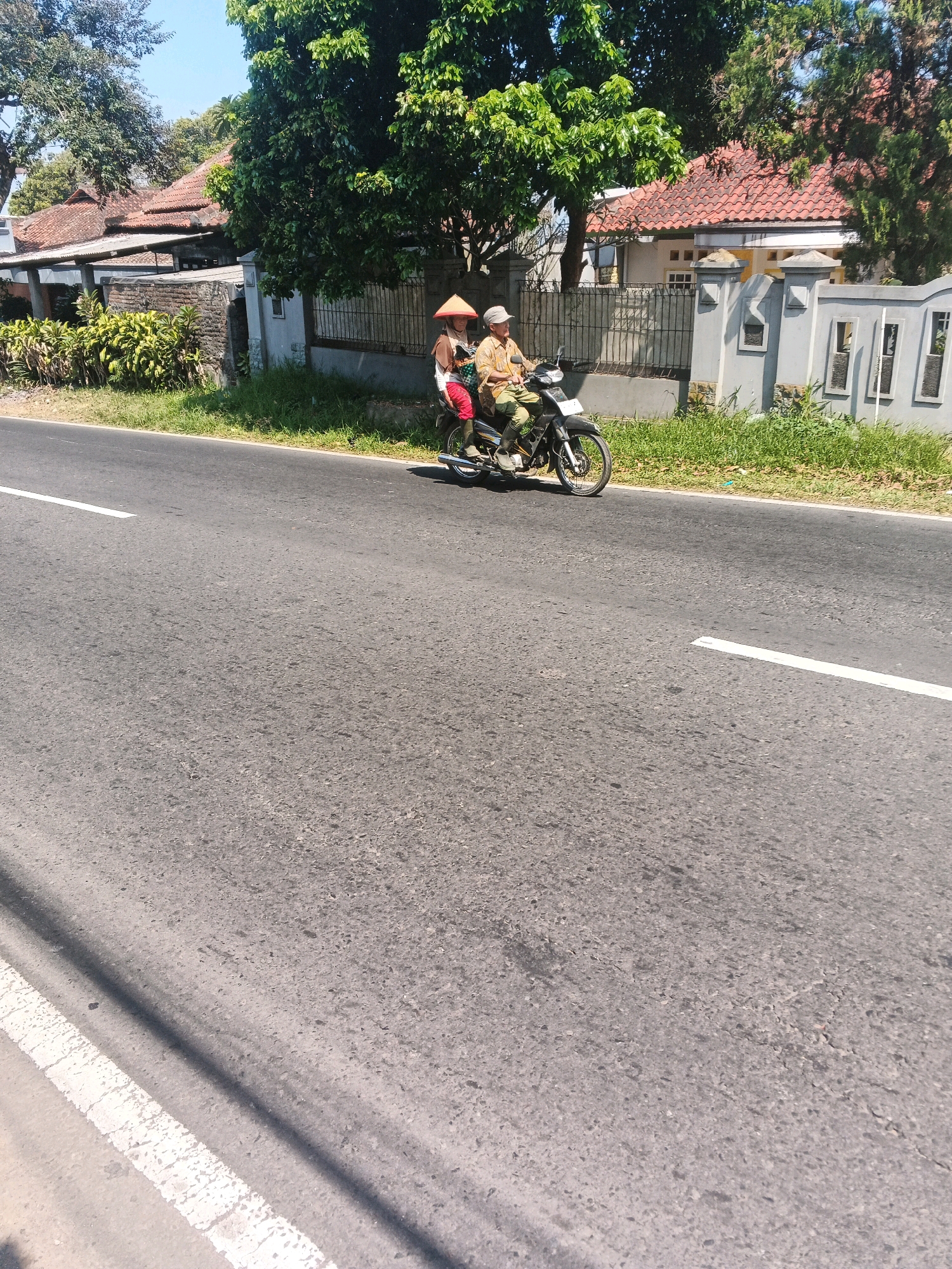 In Bandungan, Indonesia, two individuals, one driving and one as a passenger, are riding a motorcycle on a paved road. The driver, a male, is wearing a light-colored collared shirt and a light-colored cap. The passenger, a female, is wearing a conical hat, a dark-colored top, and red pants, and is seated behind the driver. They are traveling alongside a grassy area and a residential area with buildings that feature tiled roofs. The sky is clear and blue.