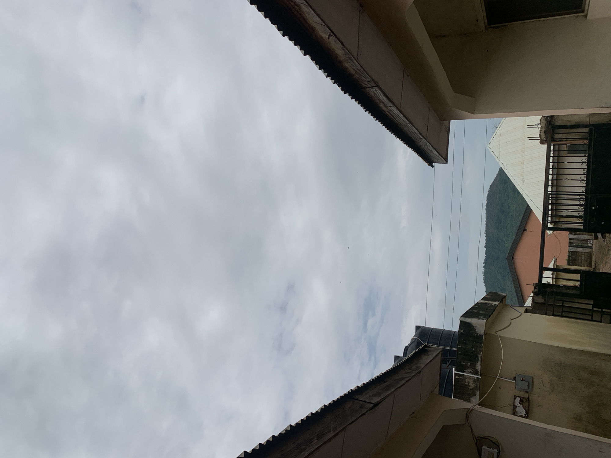 The image shows an upward view between buildings in Lokoja, Nigeria. The sky is overcast with grey and white clouds. Fragments of rooflines from adjacent structures are visible along the top and bottom edges of the frame. On the right, a building with a water tank on top is evident, alongside a metal gate and what looks like a corrugated metal roof of another structure further away. Power lines are strung across the gap between the buildings. In the background, a hill or mountain covered in greenery can be seen.
