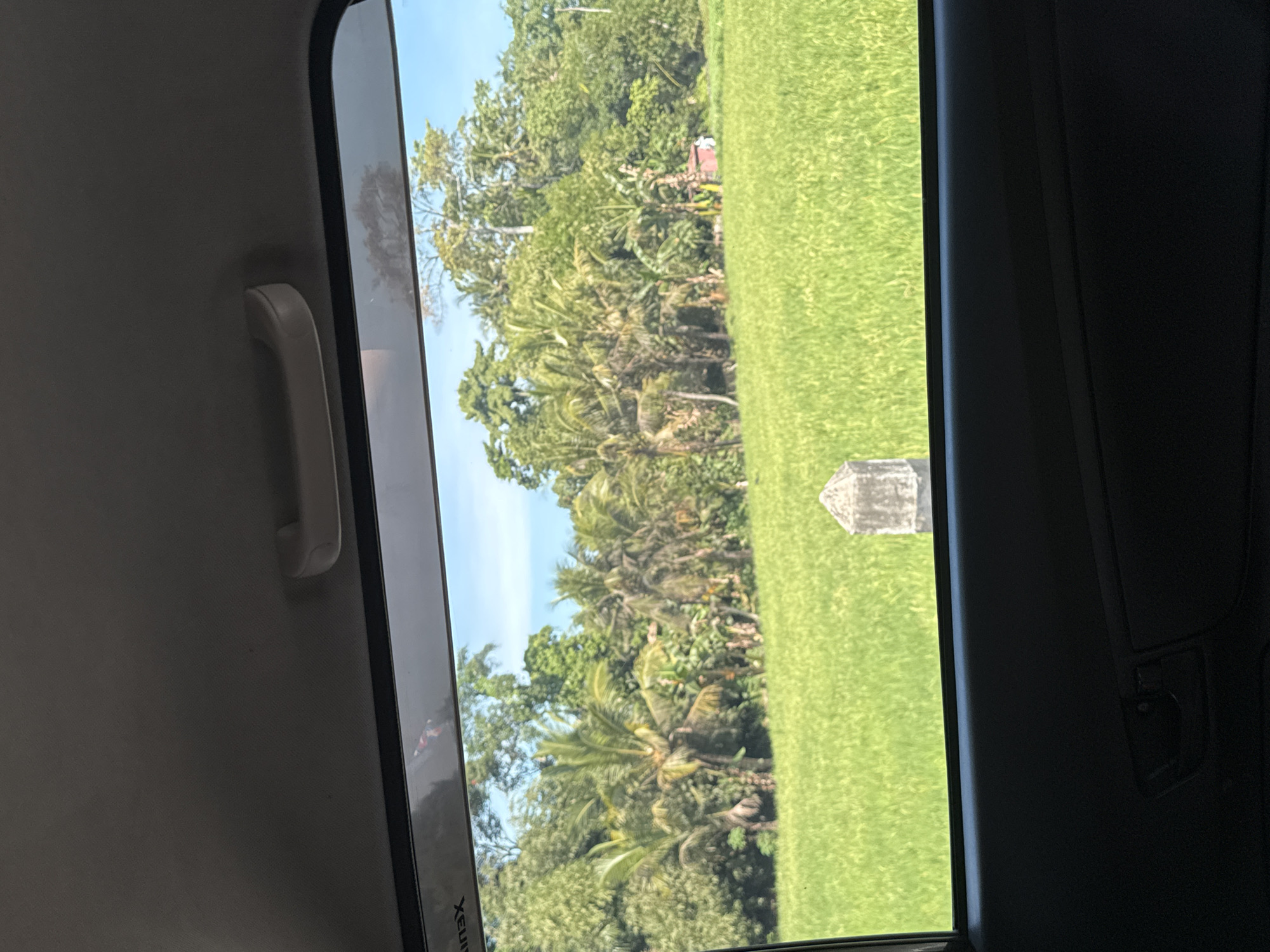 The image, taken from inside a vehicle in Sukawati, Indonesia, shows a view through the car window. In the foreground, the dark interior of the car, including the door panel and handle, is visible. The window looks out onto a vibrant green rice field under a clear blue sky. A small, weathered concrete marker stands in the field. Beyond the rice field, a lush line of tropical trees, including palm trees, forms a dense backdrop. A small red structure is partially visible among the trees. The scene suggests a rural or semi-rural landscape typical of the region.