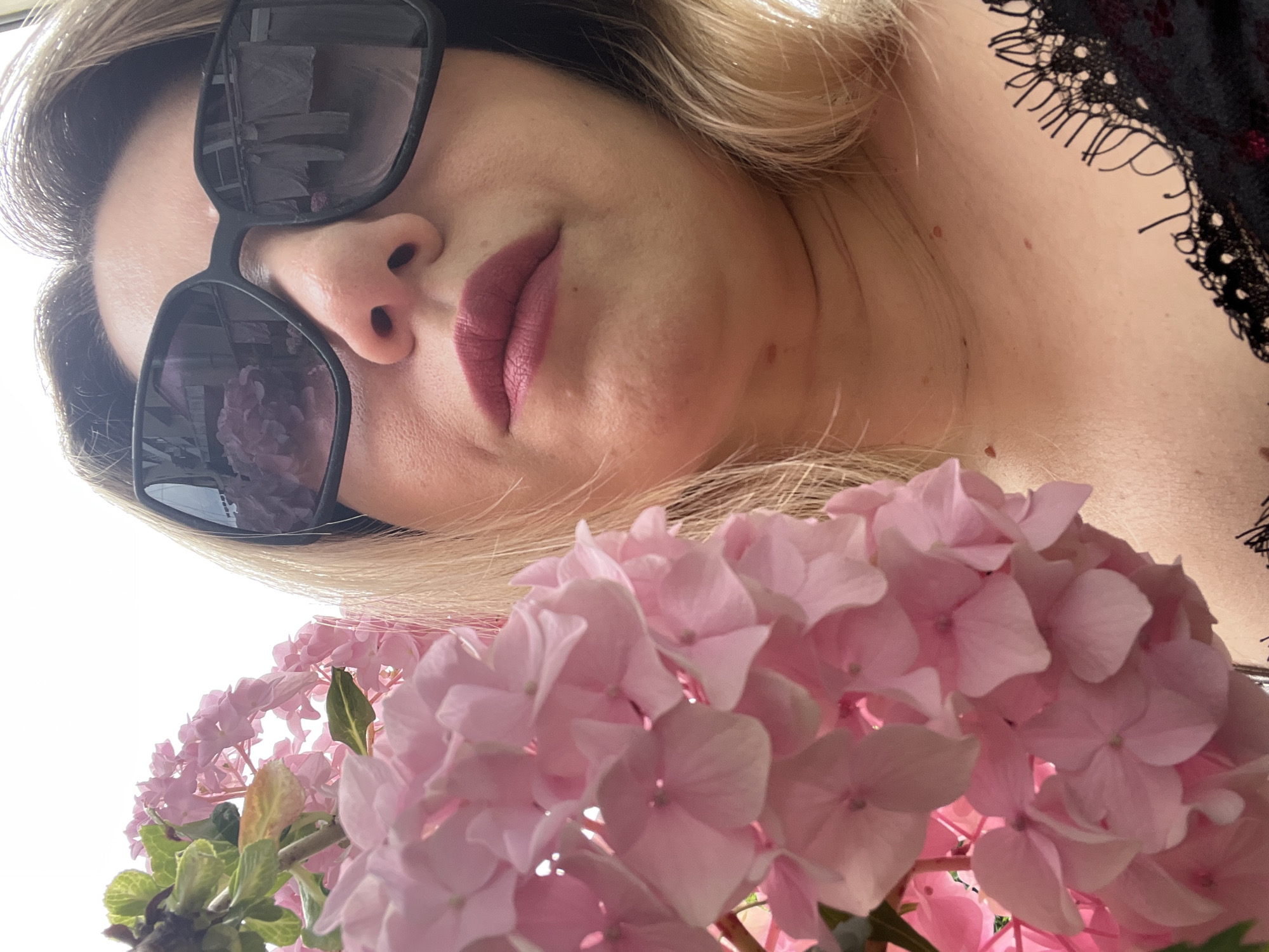 The image is a close-up, low-angle shot of a woman wearing large, black sunglasses and pink lipstick. She is holding a large cluster of pink hydrangeas in front of her face, obscuring the lower part of her face. Her blonde hair is visible around her face. In the reflection of her sunglasses, a clothesline with laundry is visible, suggesting an outdoor setting. The woman is wearing a black top with lace detailing. The image does not contain any elements that would indicate it was taken in Sochi, Russia.